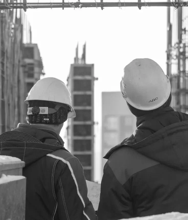 Two construction workers wearing helmets and jackets, looking at a construction site with buildings and scaffolding.