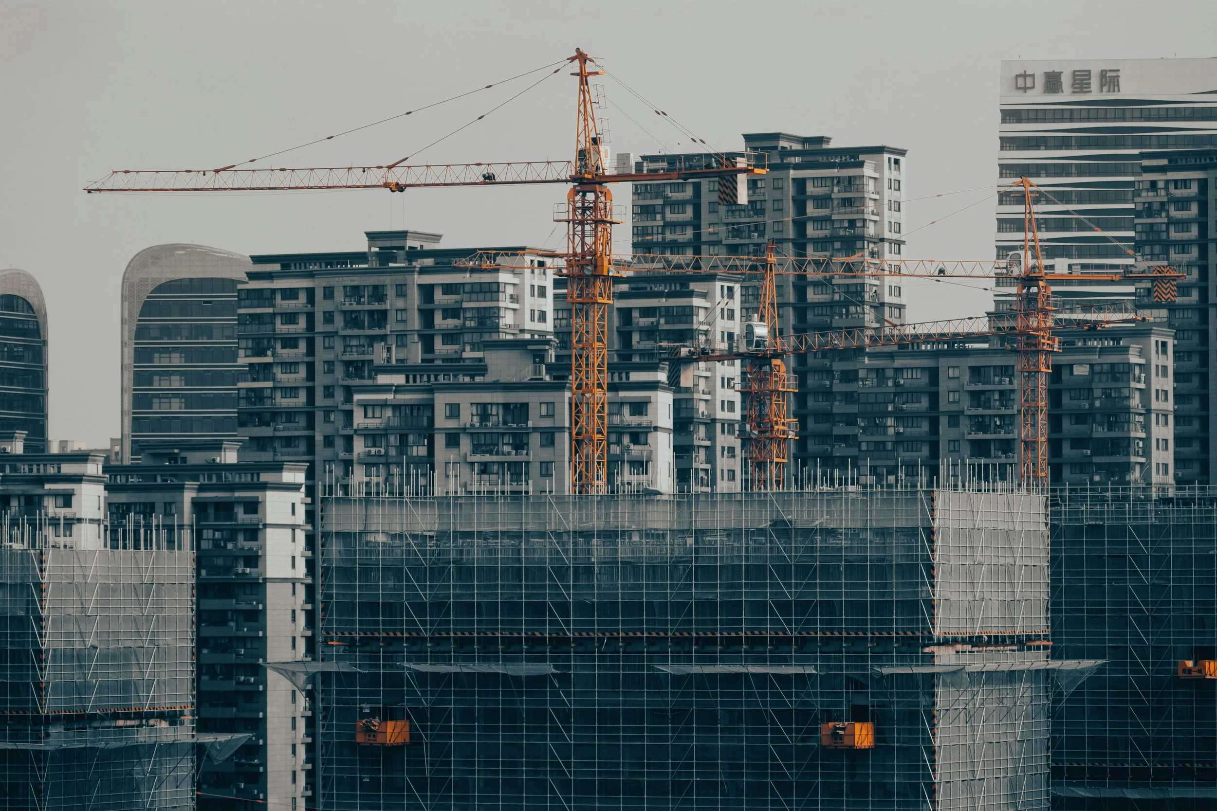 Residential tower blocks under construction with orange cranes and scaffolding against an overcast sky.