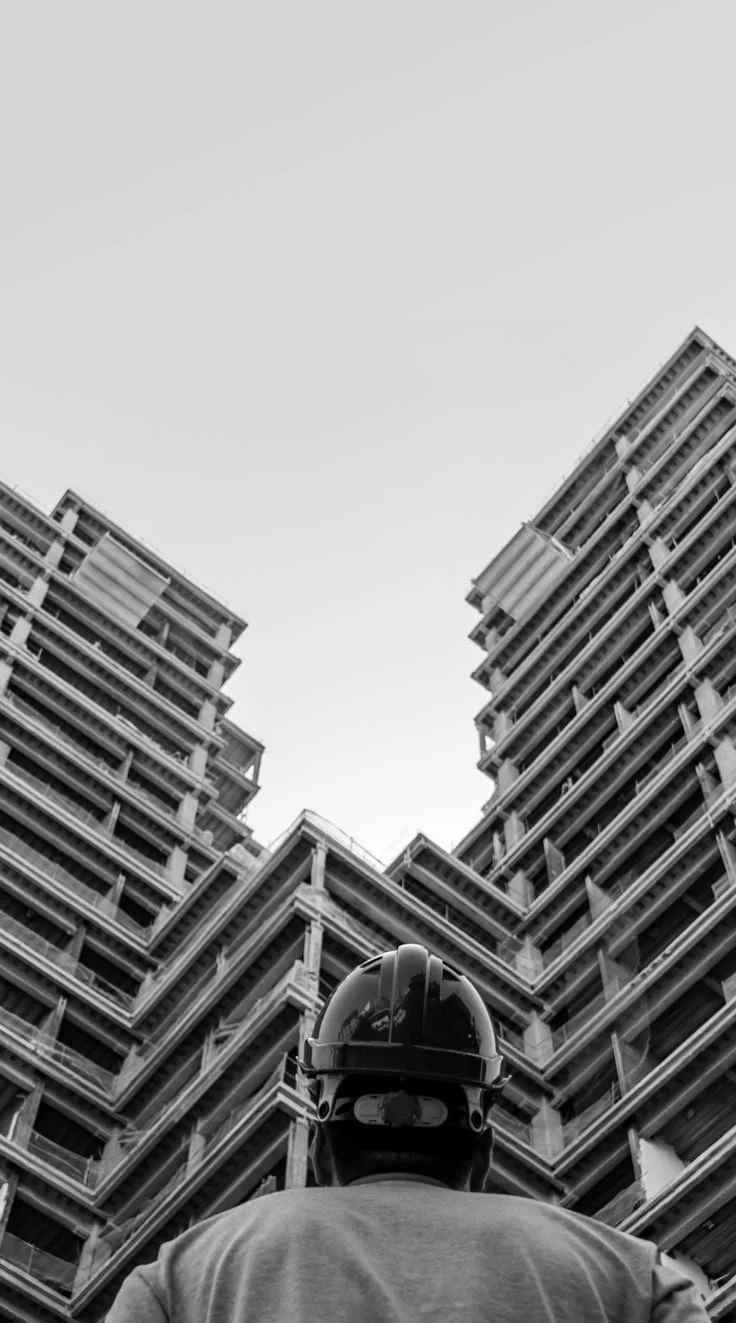 A construction worker wearing a helmet looking up at tall, multi-story buildings under construction.
