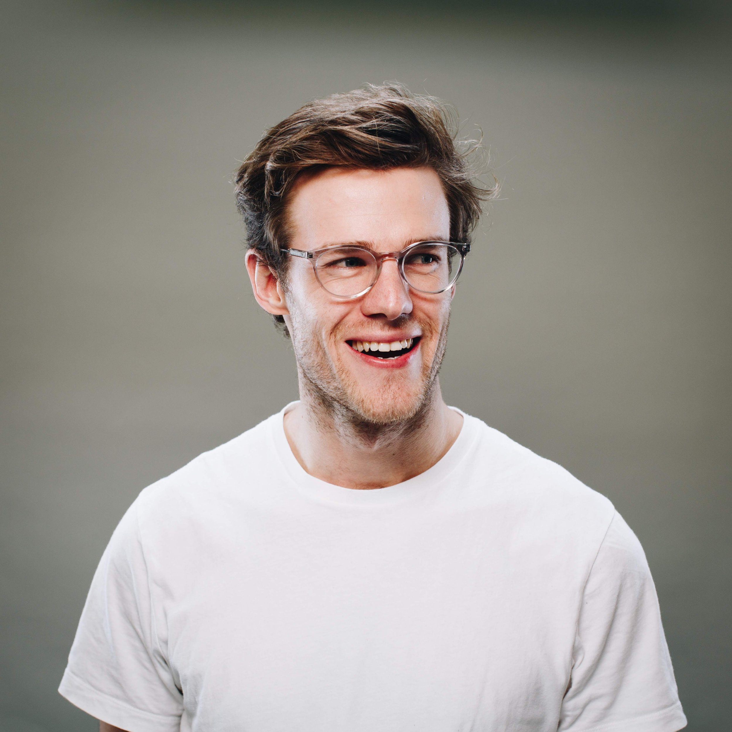 Young man with light skin, glasses, and brown hair smiling and looking to the side, wearing a white t-shirt, against a gray background.