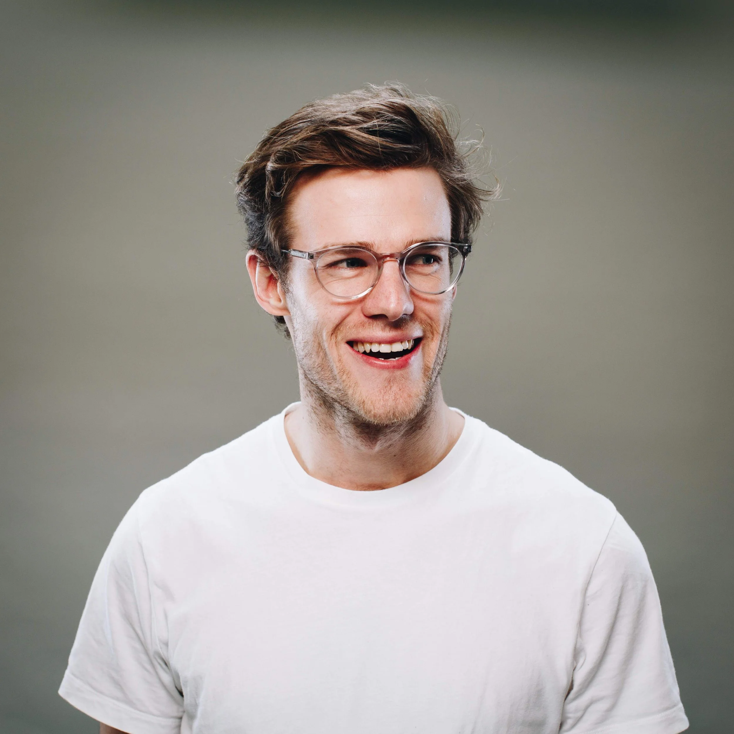 Smiling young man with glasses and white t-shirt, looking to his right, against a plain green background.