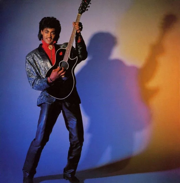 A person dressed in 1980s style clothing holding an acoustic guitar in front of a colorful, gradient background.