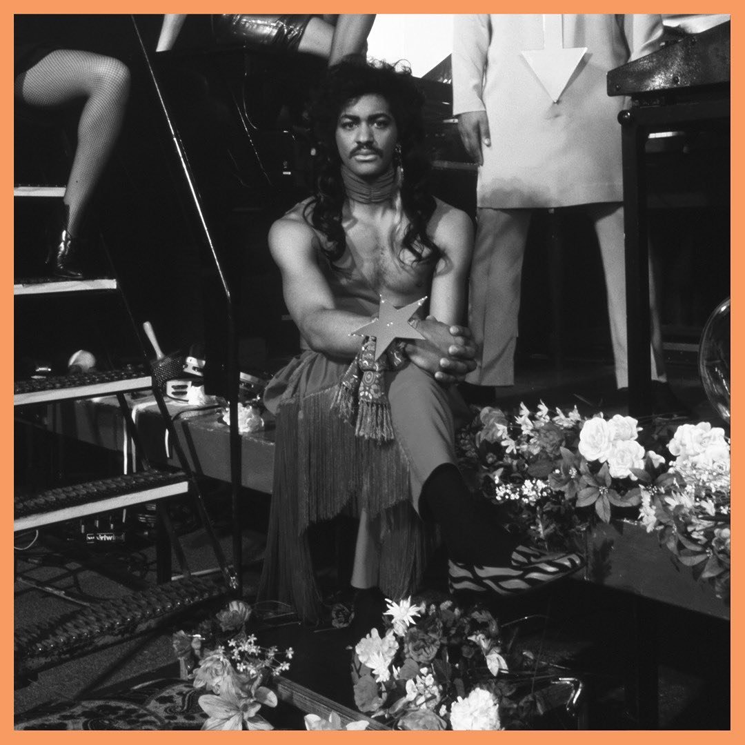 with long, dark, curly hair and a mustache, sitting in a room decorated with flowers, holding a star-shaped award. They are dressed in eclectic clothing, with a fringed skirt and patterned footwear, surrounded by various objects and another person partially visible in the background.