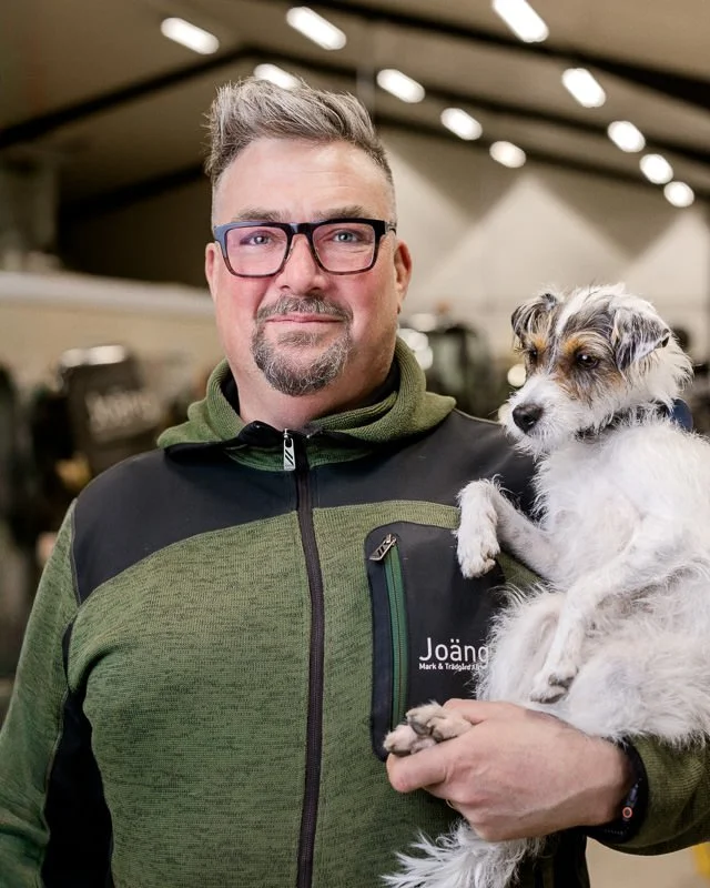 Man håller en liten hund i en inomhusmiljö, båda tittar mot kameran.