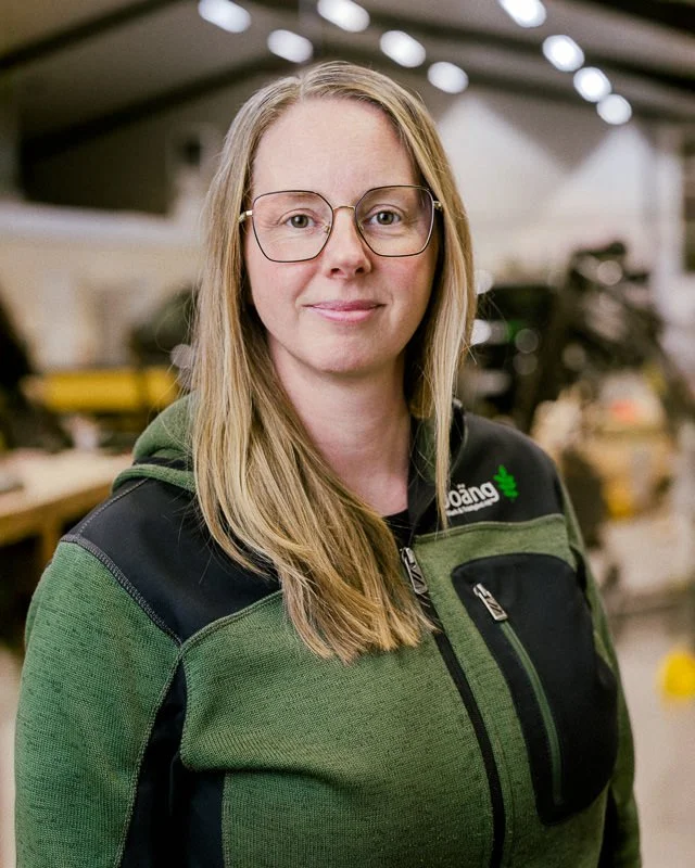 En kvinna med glasögon och lång blont hår, klädd i en grön och svart arbetsjacka, står i en verkstad eller fabriksok, bakgrund med verktyg och maskiner.