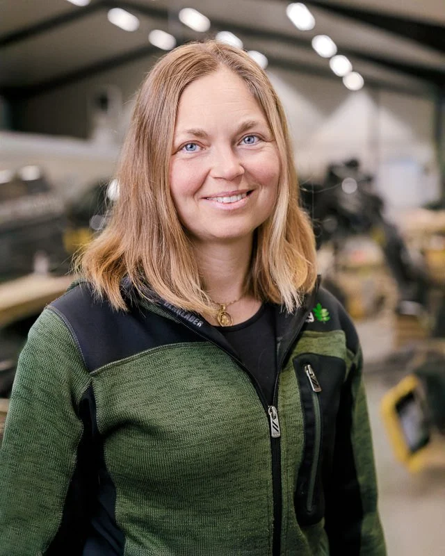 Kvinna med ljus brunt hår i en grön och svart jacka, står i en verkstad.
