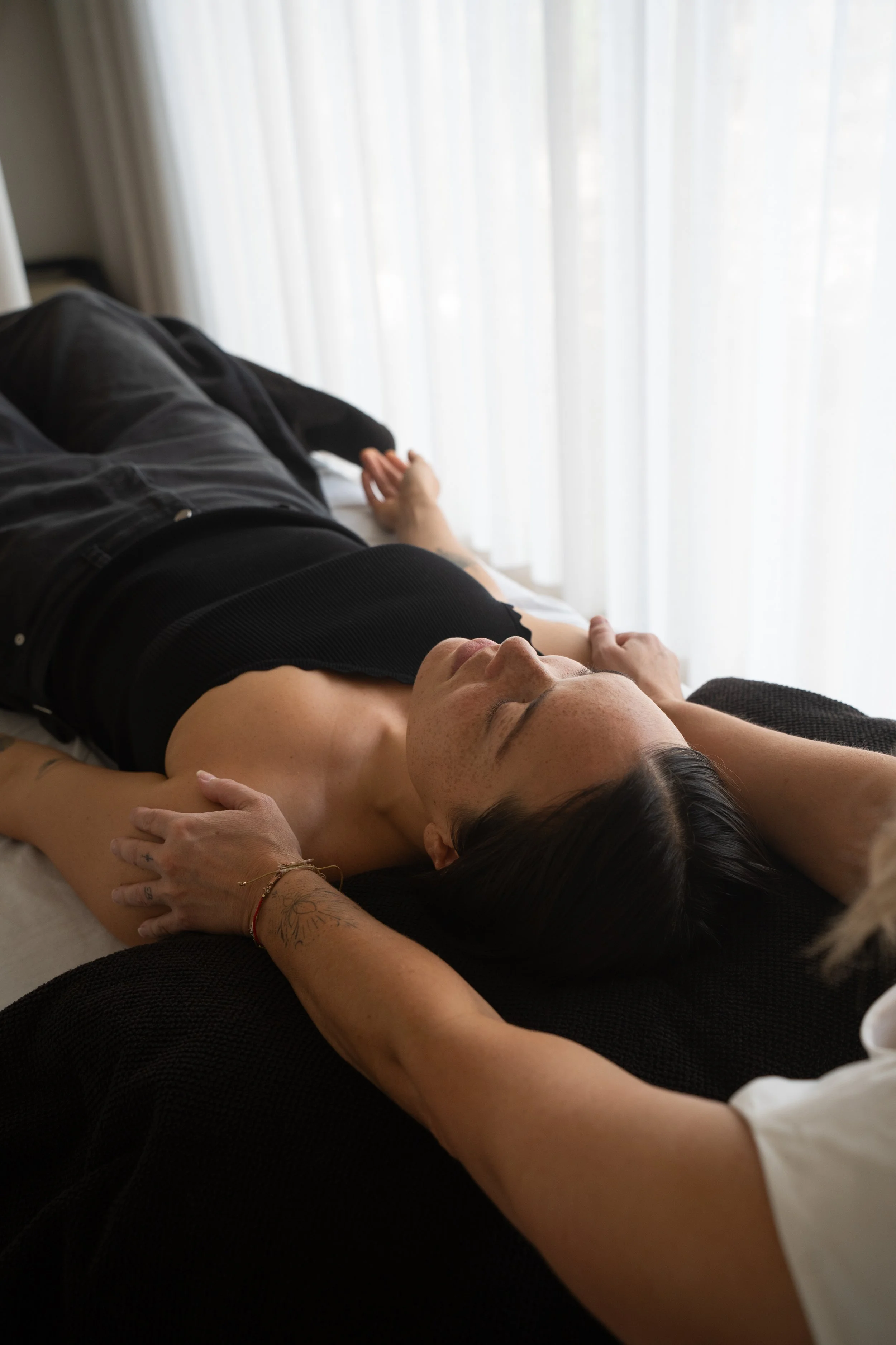 A person lying on a bed with their eyes closed, arms stretched out, and a hand resting on their stomach, next to a nightstand with a black lamp. She is wearing a black sleeveless top and black pants, with a bracelet on her wrist.