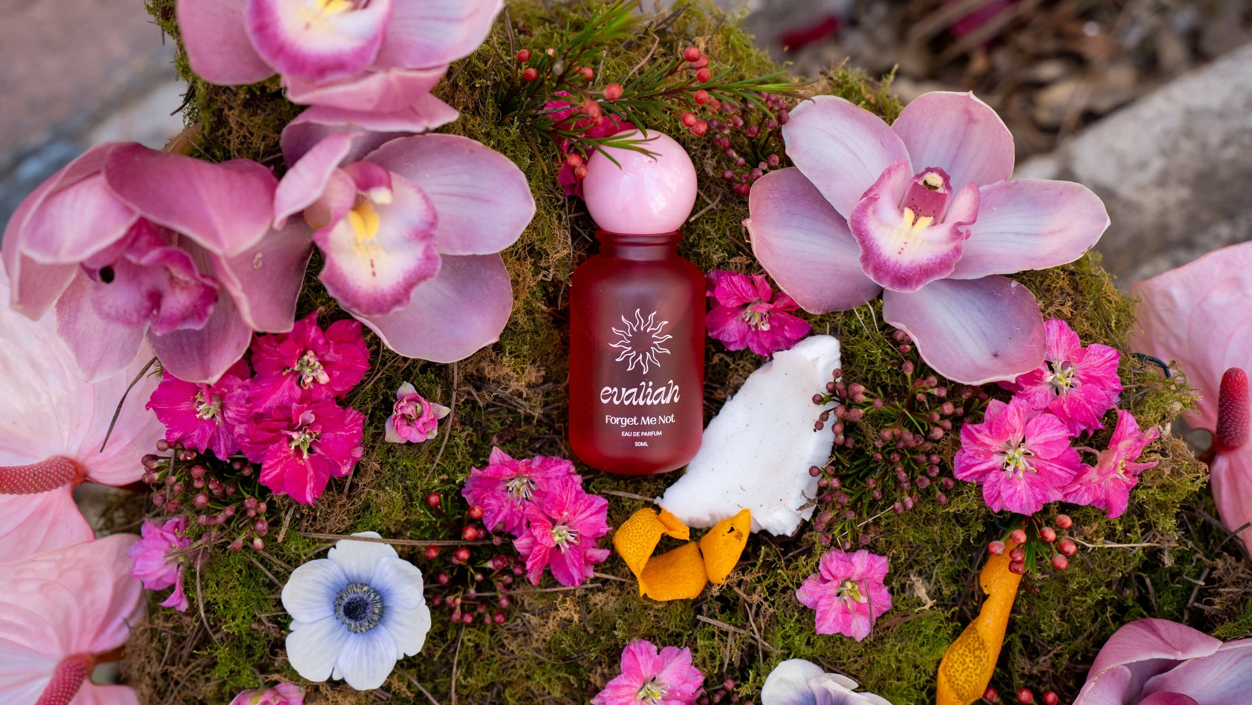 A bottle of Evaliah perfume called 'Forget Me Not' placed among pink orchids, moss, and small pink flowers on a natural surface.