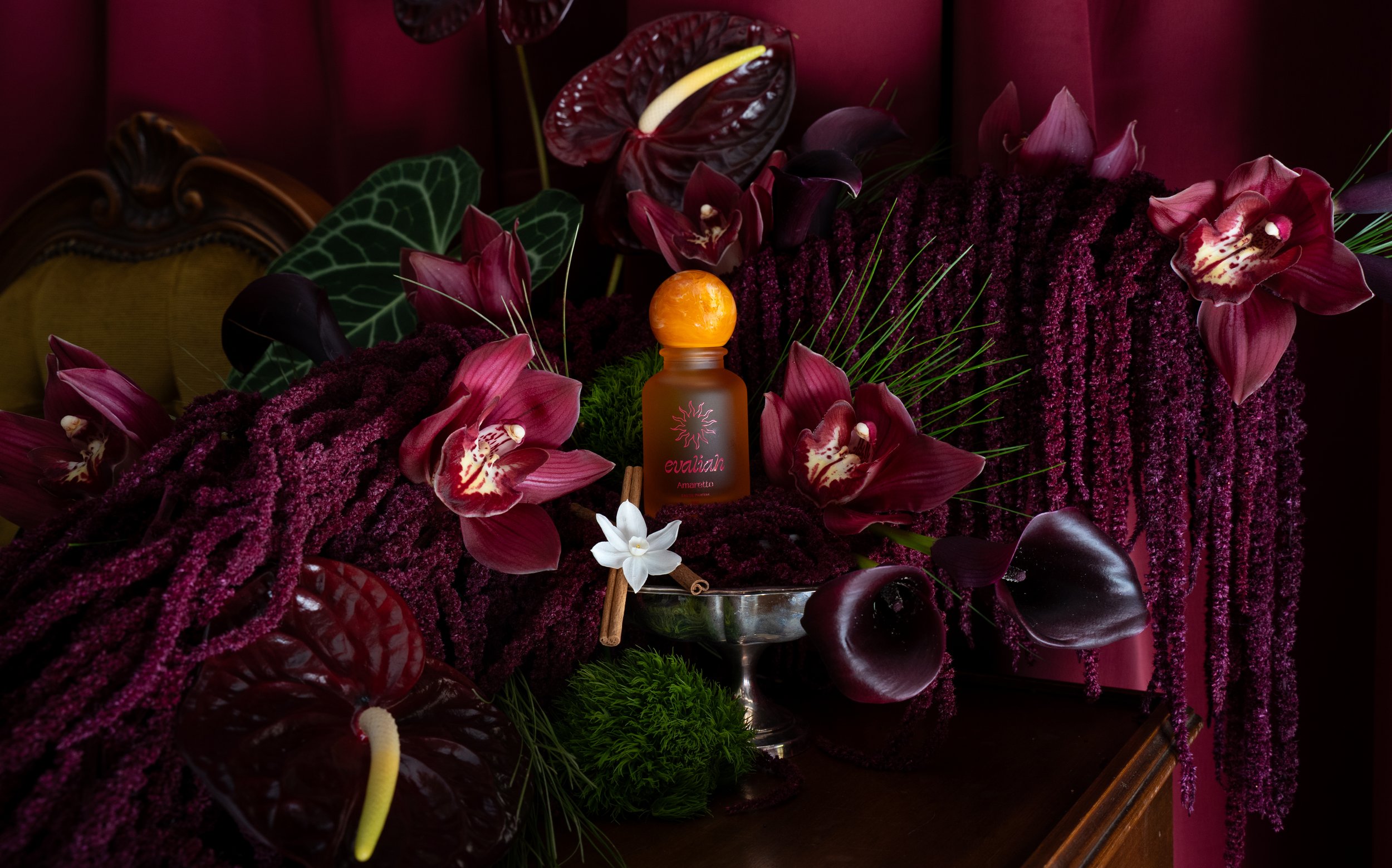 A decorative floral arrangement with burgundy, purple, green, and white flowers, including orchids and anthuriums, surrounding a perfume bottle labeled 'evahh Amaretto' on a table with a dark background.
