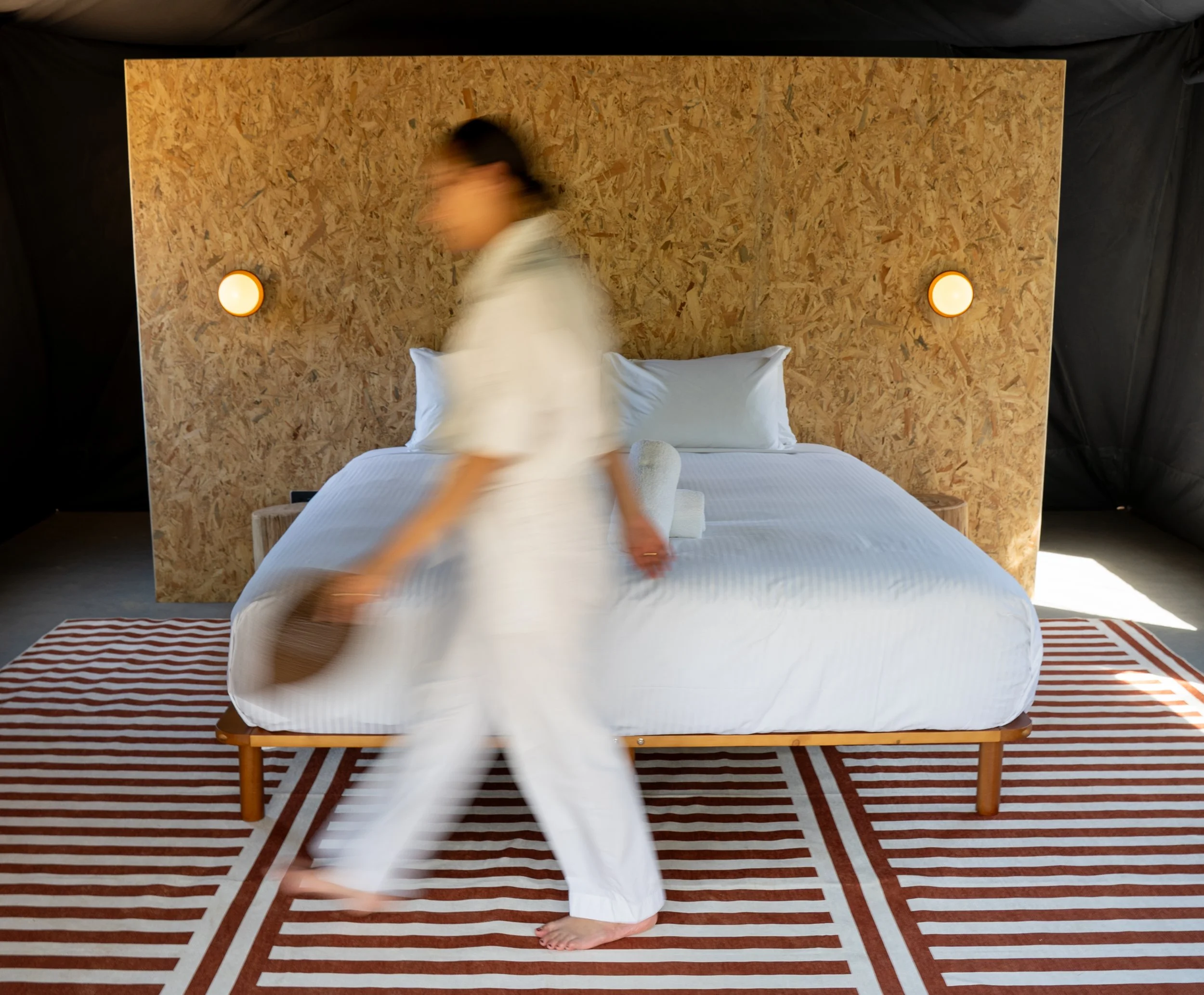 A person walking across a bed with white bedding, levitating or jumping, in a room with a wooden headboard, two wall-mounted circular lights, a striped rug, and a black backdrop surrounding the scene.