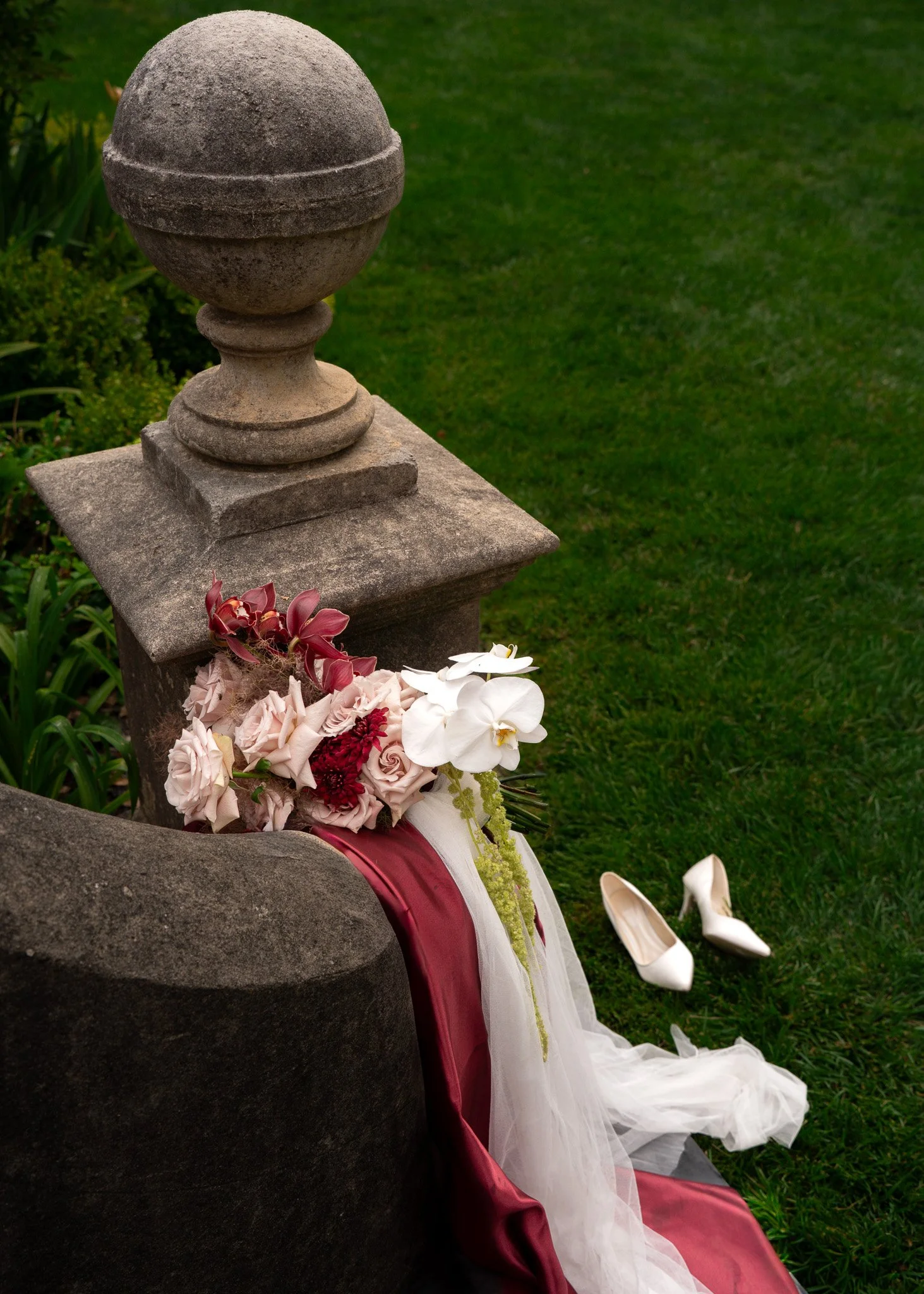 A stone grave marker in a cemetery with a floral arrangement and white high heels nearby on a grassy area.