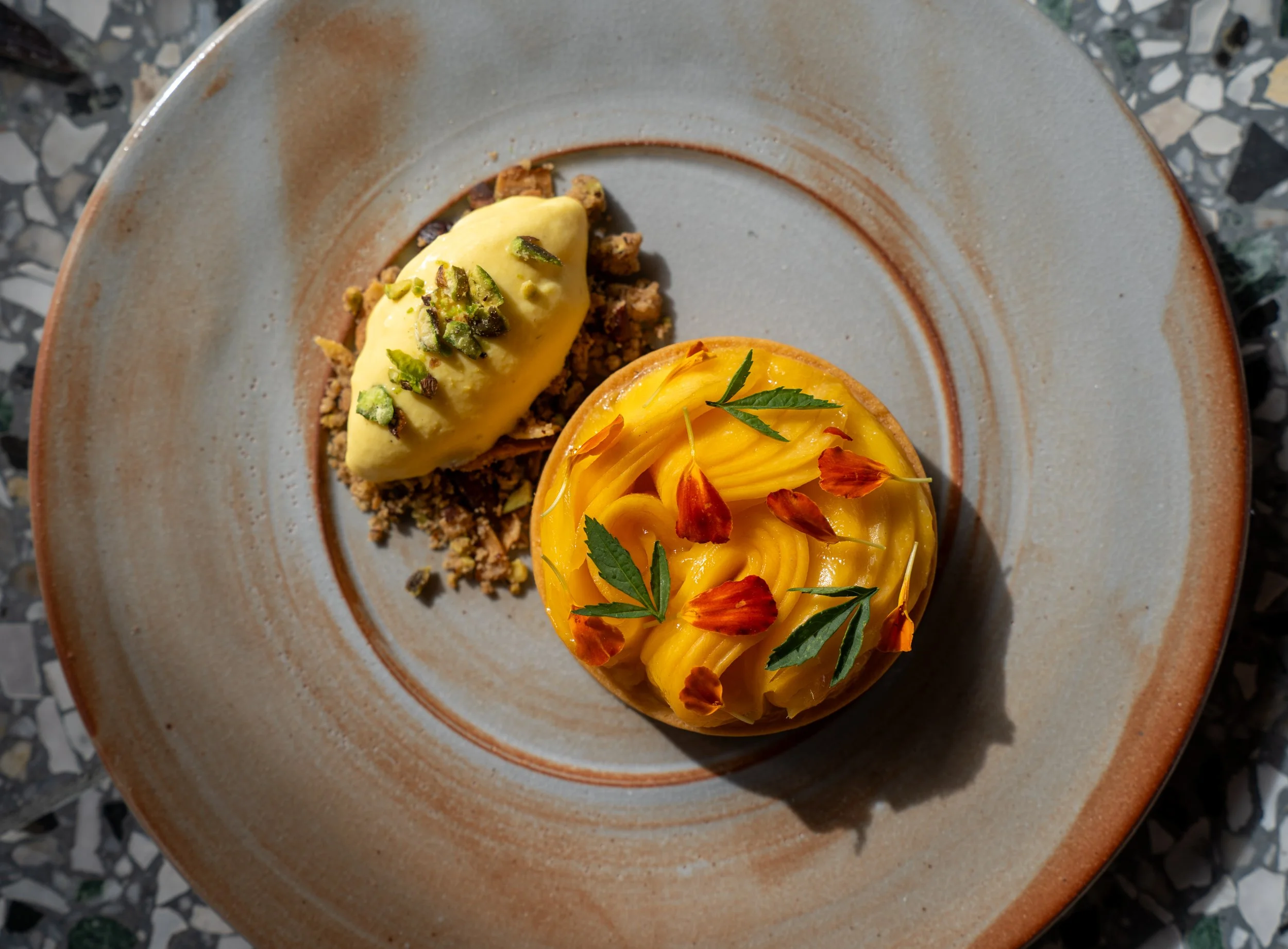 Dessert plate with two types of mousse: one yellow topped with edible flower petals and green leaves, and the other green topped with chopped pistachios, served on a ceramic plate.