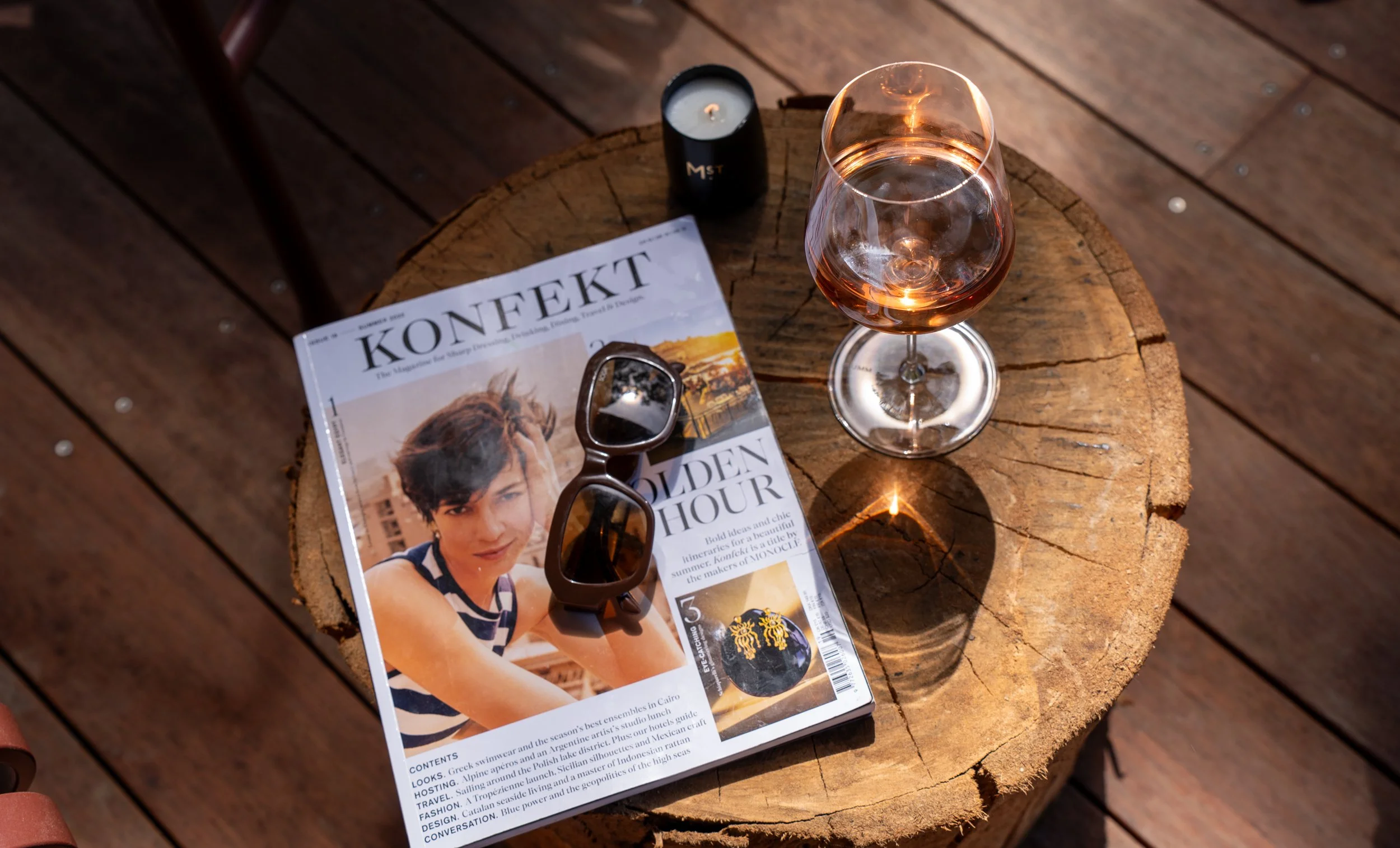 A glass of rosé wine, a magazine titled 'KONFEKT', a pair of sunglasses, a small candle, and a lighter on a wooden surface.