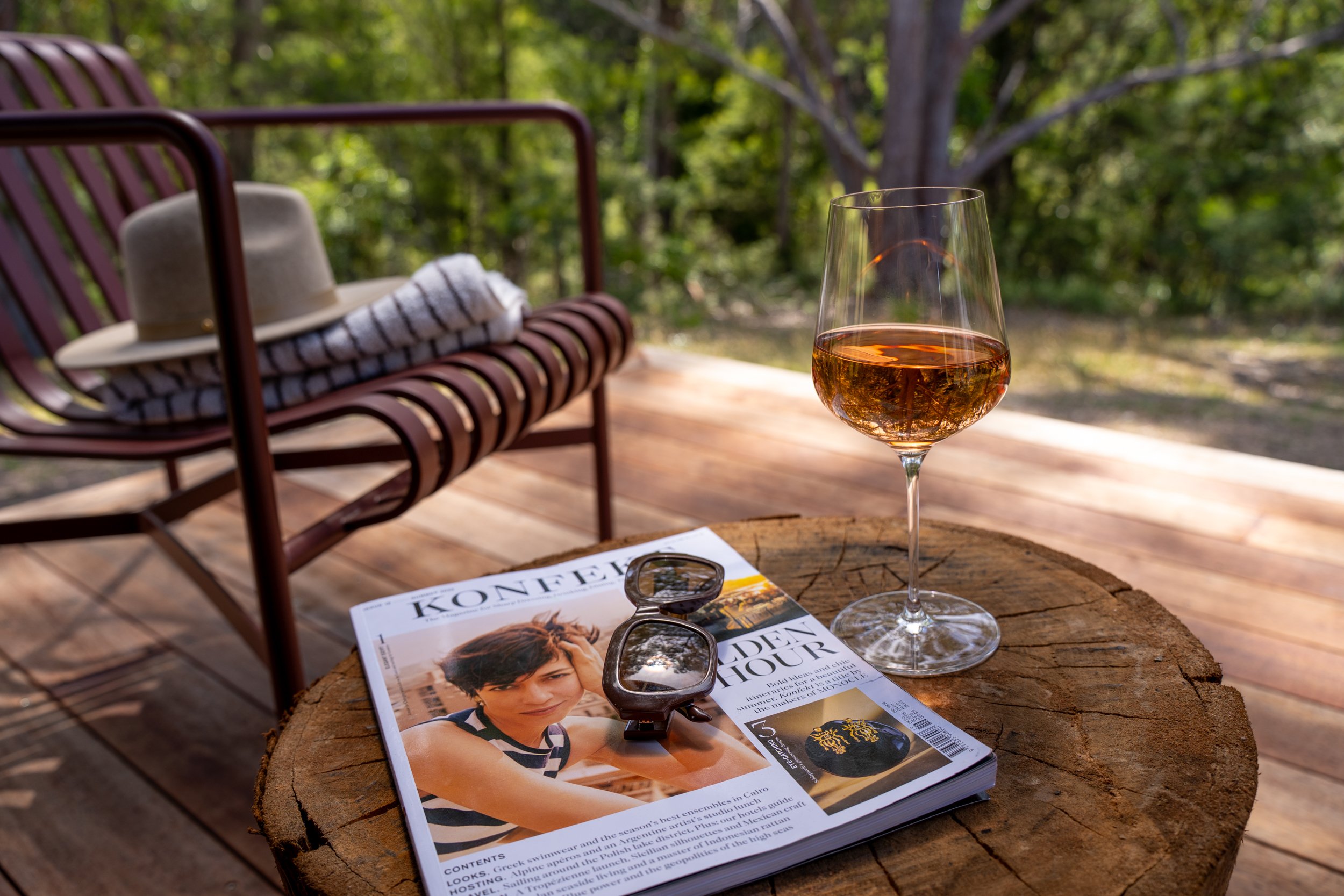 A glass of rosé wine on a wooden table outdoors with a magazine, sunglasses, a hat, and a towel on a bench in the background surrounded by trees.