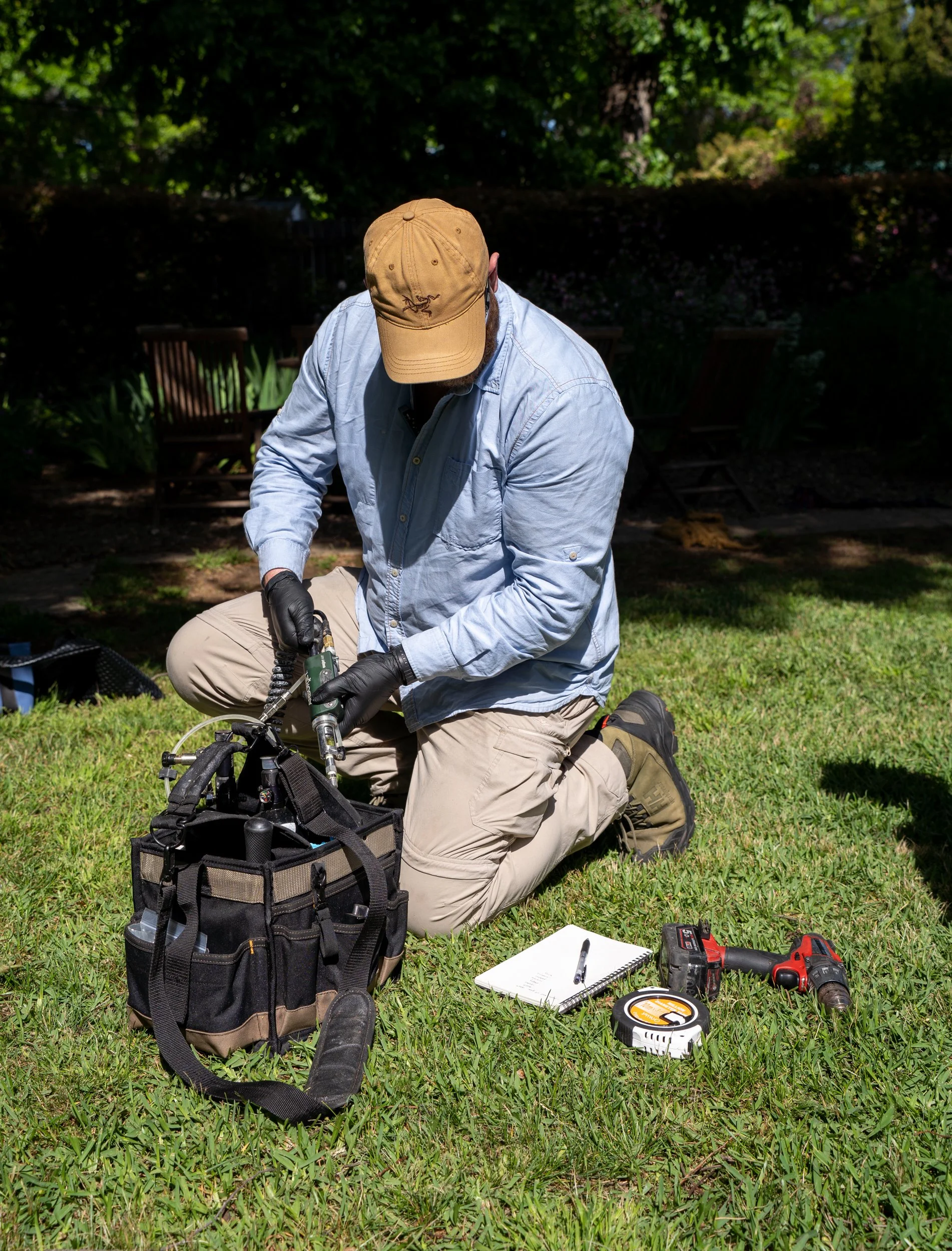 A man kneeling on the grass outdoors, wearing a tan cap, light blue shirt, and khaki pants, working with tools around him. There are chairs, a backpack, a notebook, a pen, a measuring tape, and power tools nearby, with trees and shrubbery in the background.