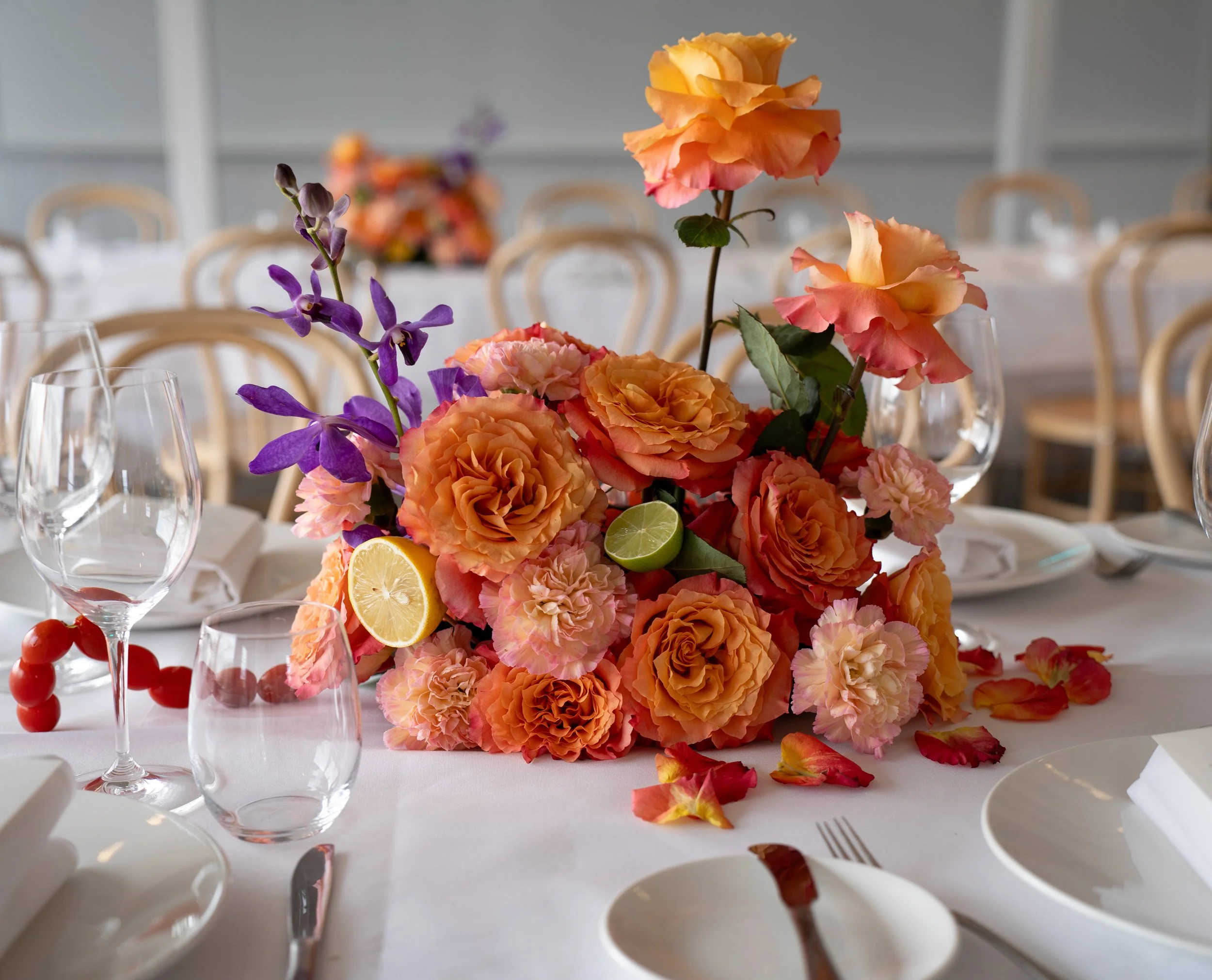 A floral centerpiece with orange, pink, and purple flowers, including roses and orchids, on a white tablecloth surrounded by empty wine glasses and scattered rose petals, set for a formal event with wooden chairs in the background.