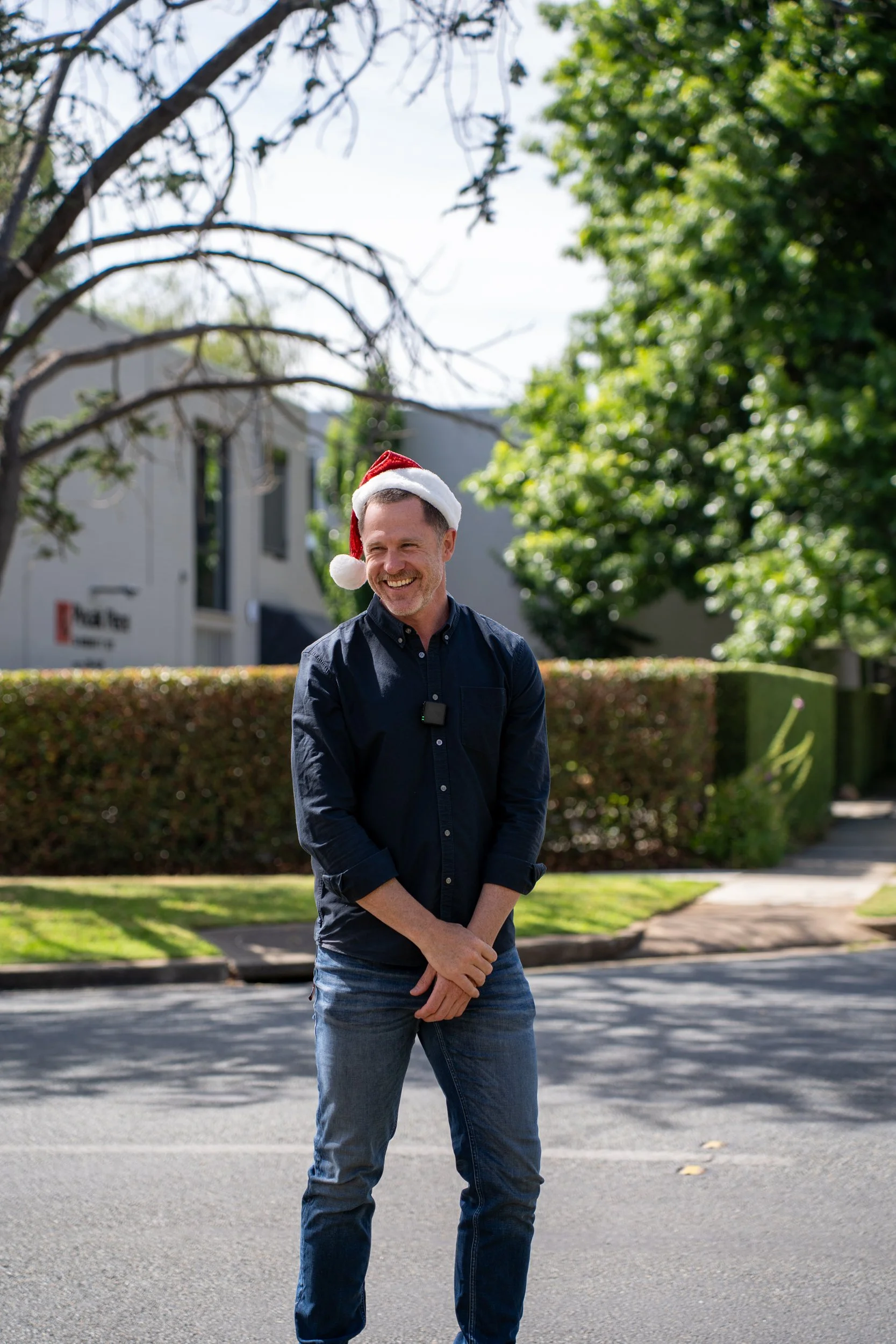 A man wearing a Santa hat and casual clothes standing outdoors on a sunny day with trees and buildings in the background.