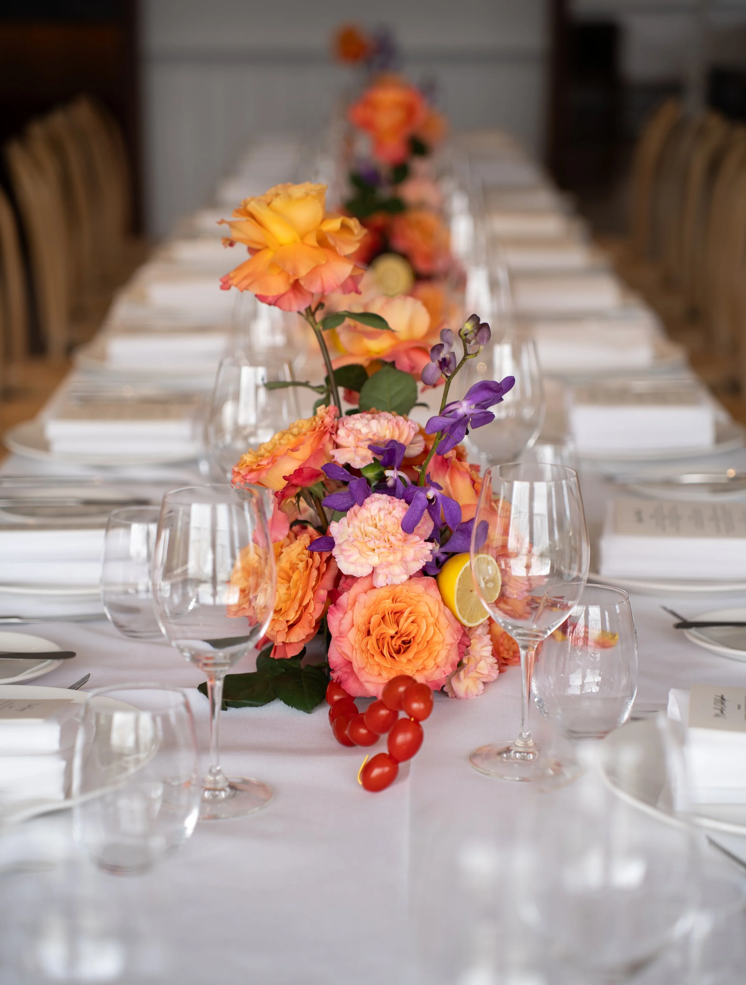 A long banquet table decorated with a vibrant floral centerpiece, surrounded by empty wine glasses and white napkins, set for a formal event or dinner.