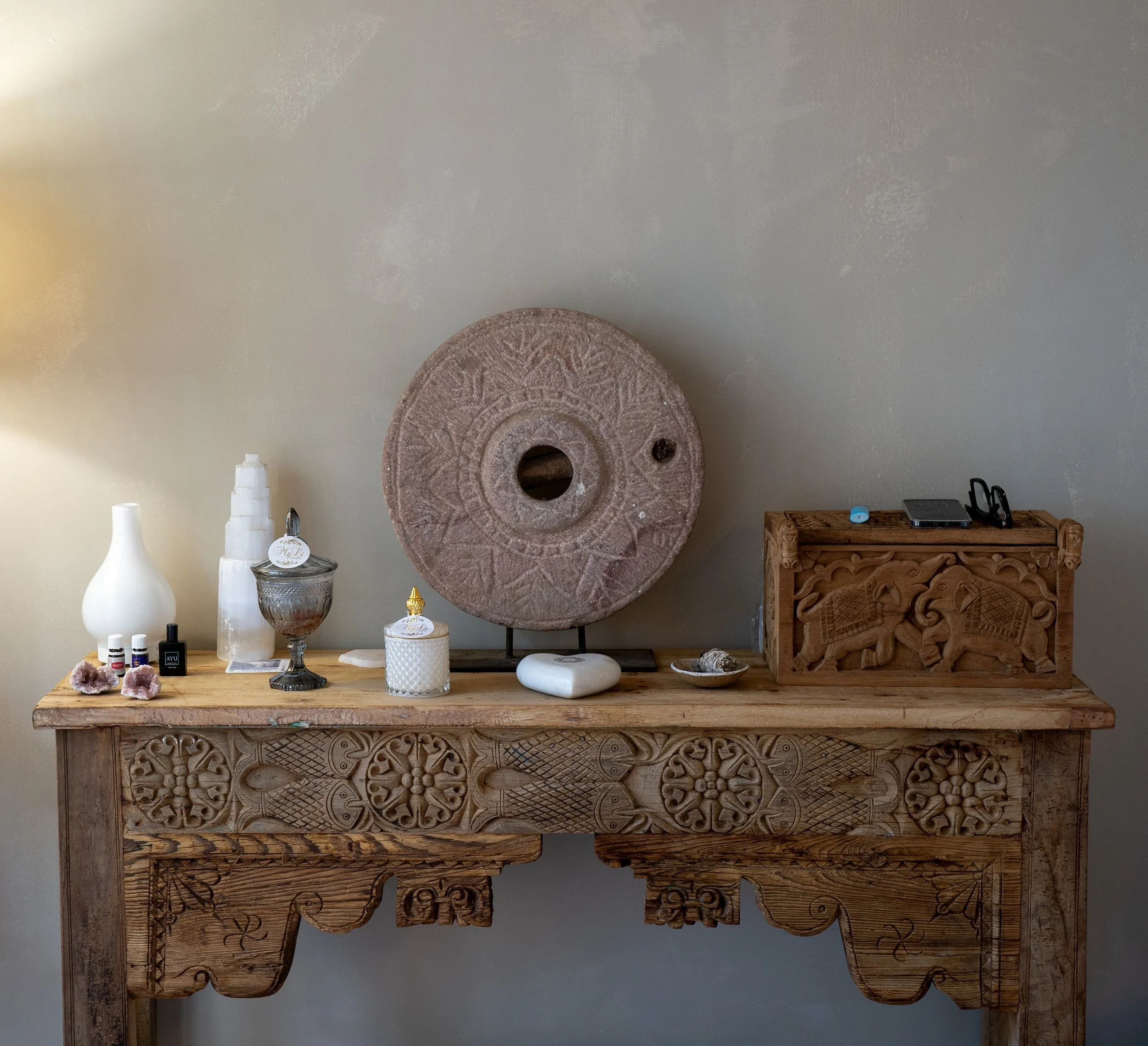 A wooden table displaying various decorative items including white vases, glassware, crystals, and small containers, with a carved wooden panel or artwork hanging on the wall behind the table.