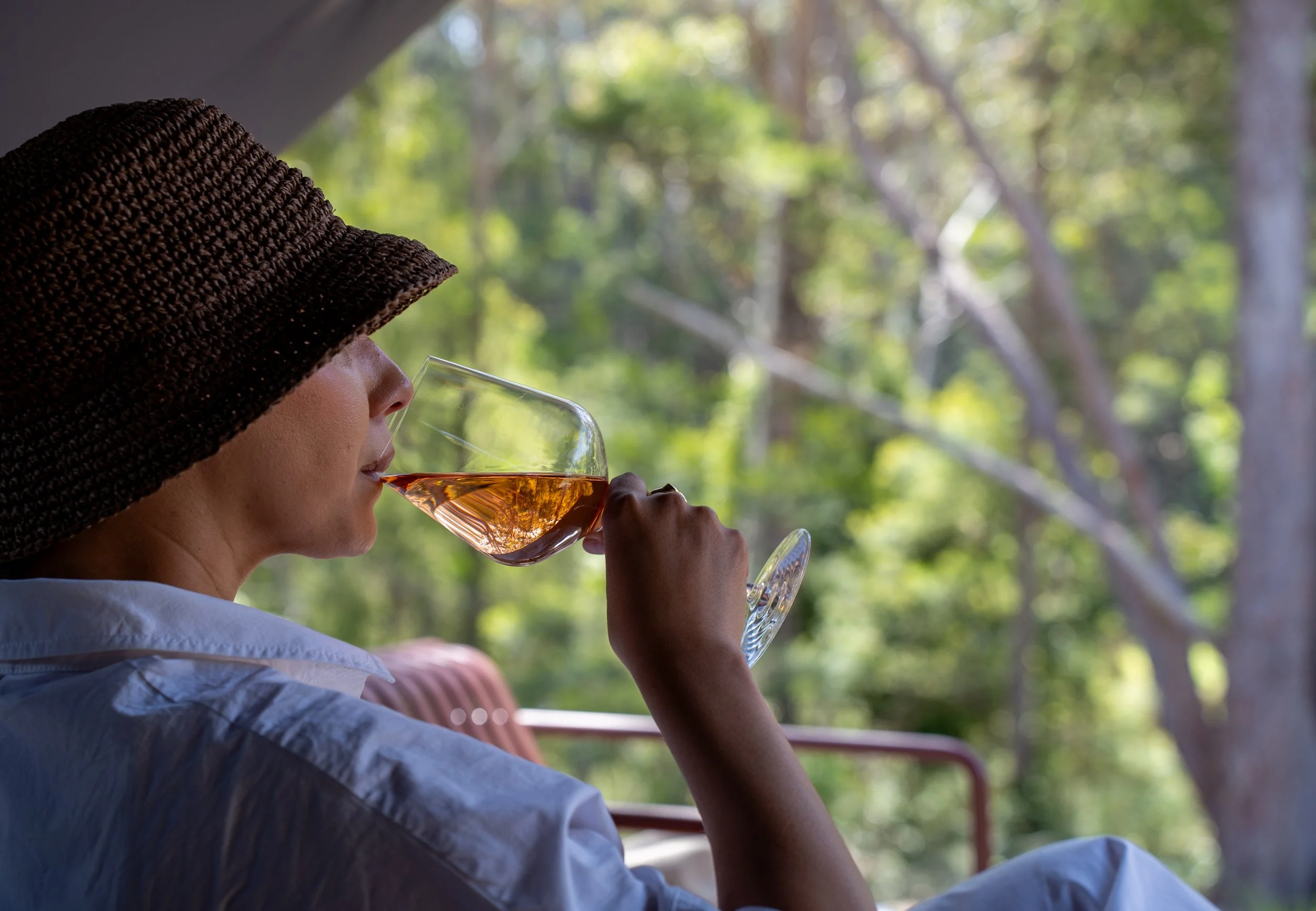 A woman wearing a large woven sunhat drinking rosé wine from a glass outdoors with a blurred green forest background.