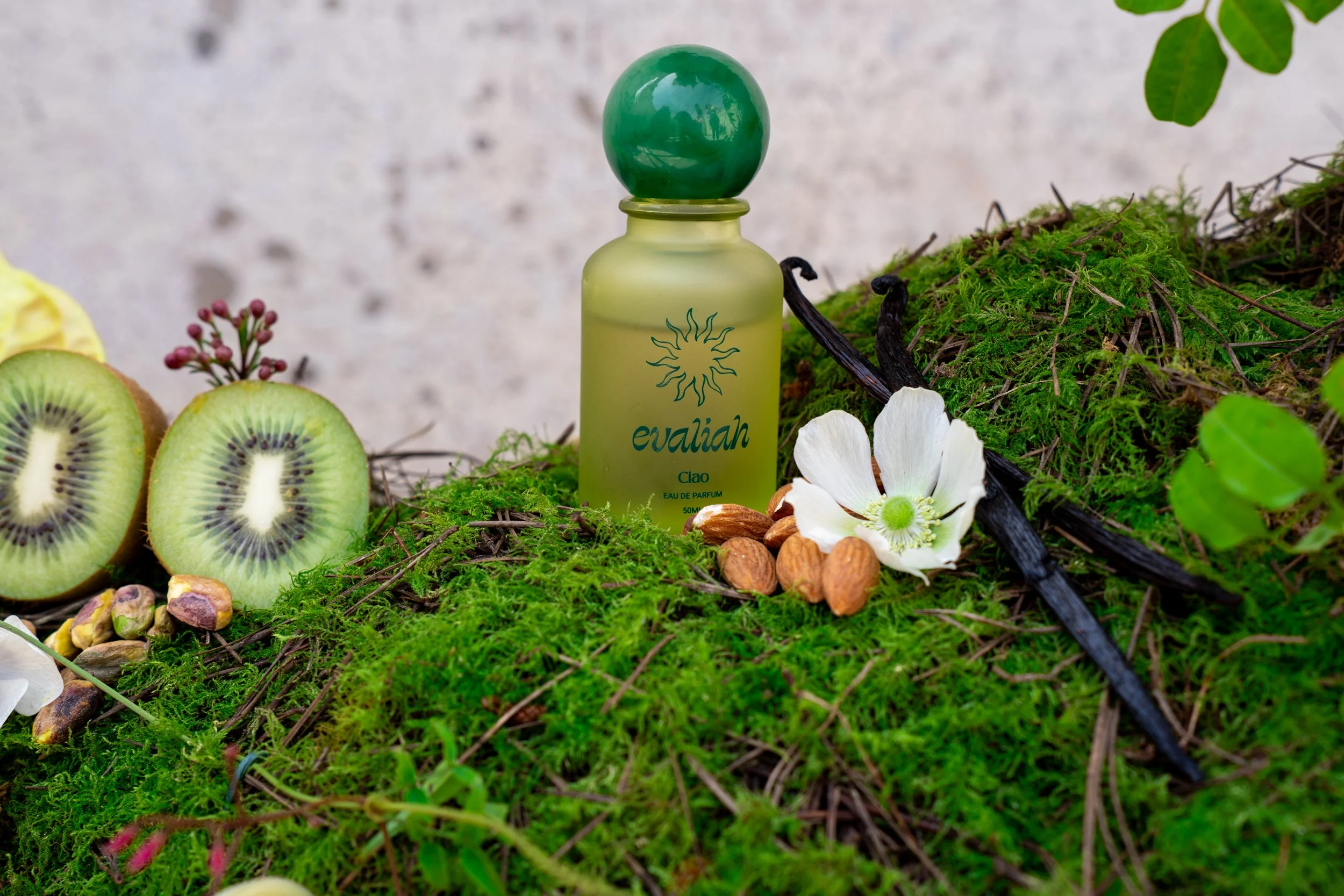 A bottle of Evaliah perfume labeled 'Ciao' placed on mossy greenery surrounded by sliced kiwi, almonds, white flowers, and sprigs of pink and purple berries, with a gray background.