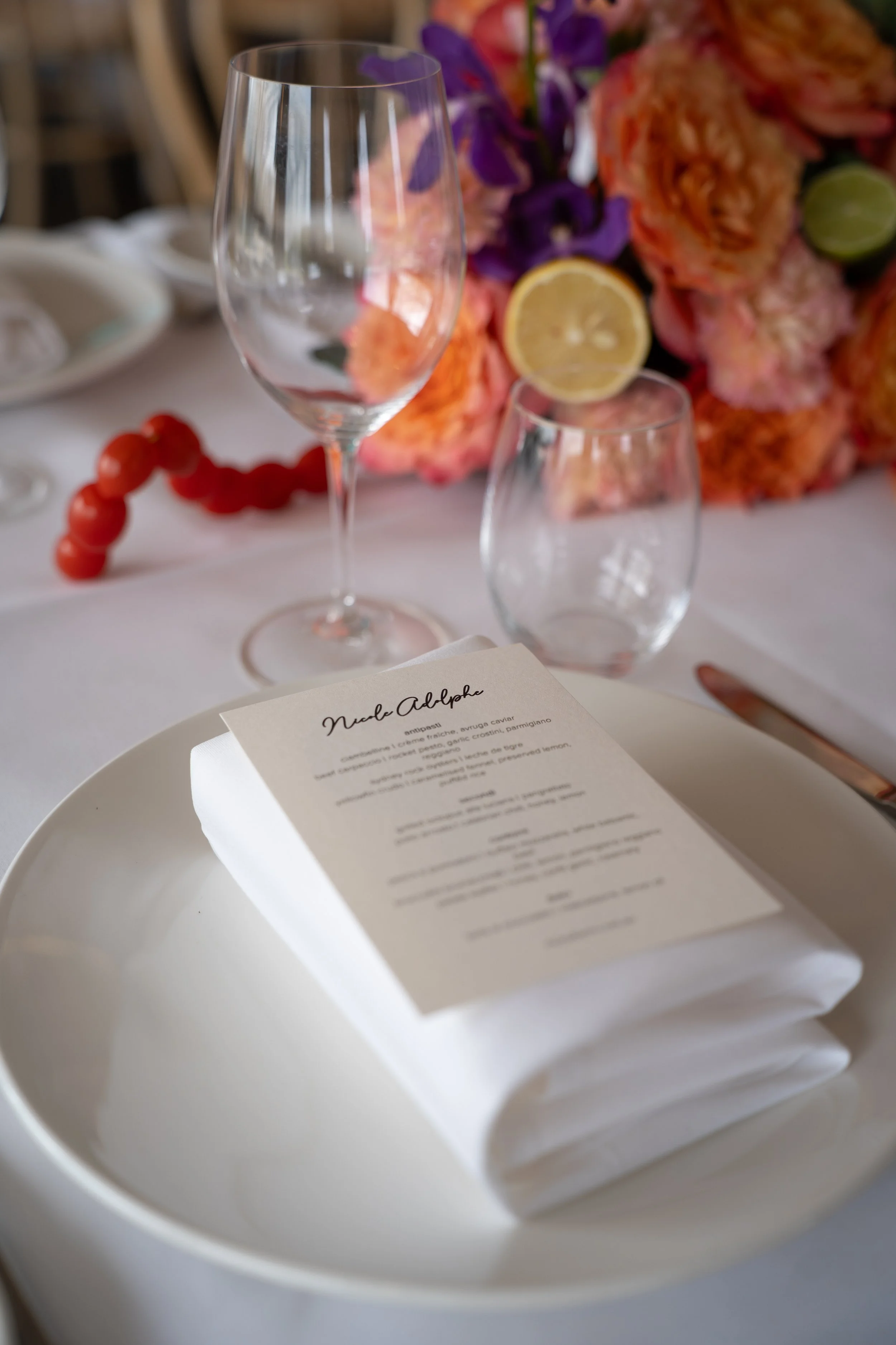Empty wine glass, partially filled glass with lemon slice, floral arrangement in the background, white tablecloth, menu on a white plate in the foreground.