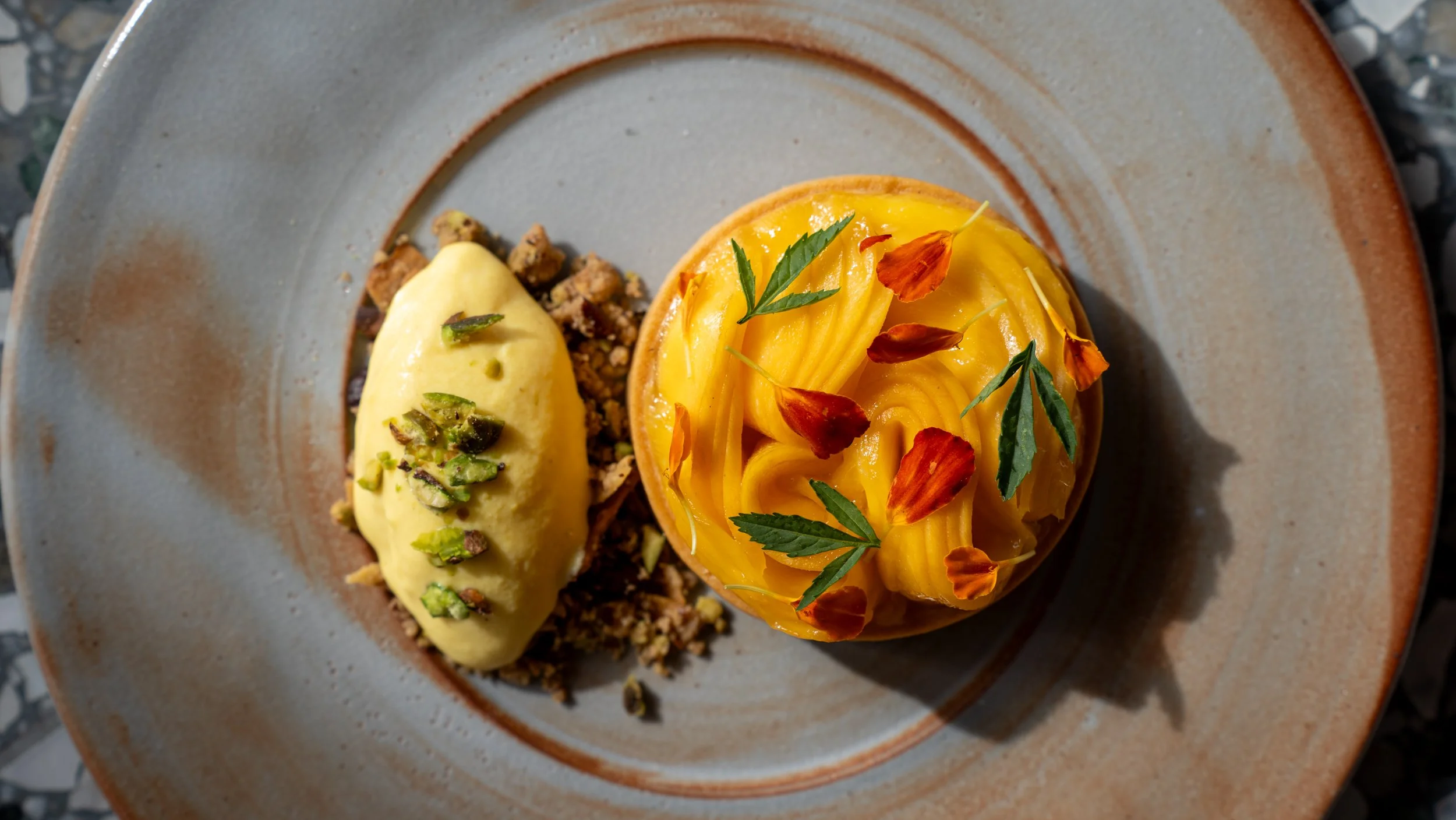A dessert with yellow gel-like topping decorated with green leaves and orange edible flowers on a round pastry base on a plate.