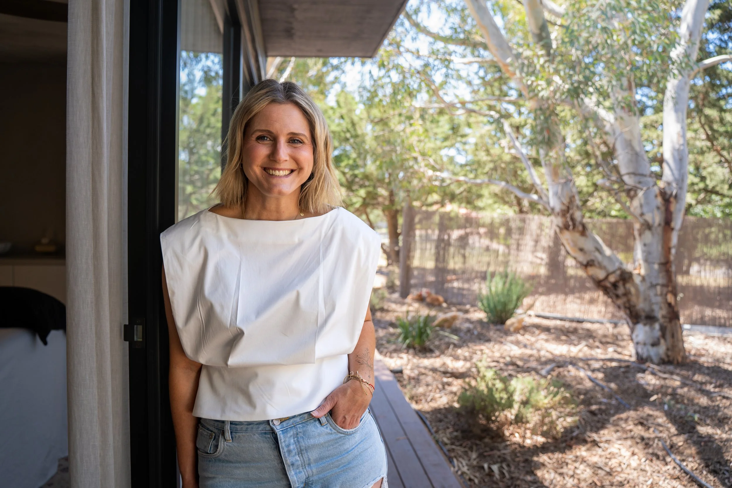 A smiling woman with blonde hair standing near a sliding glass door, wearing a white top and blue jeans, with a backyard visible outside featuring trees and a wooden fence.