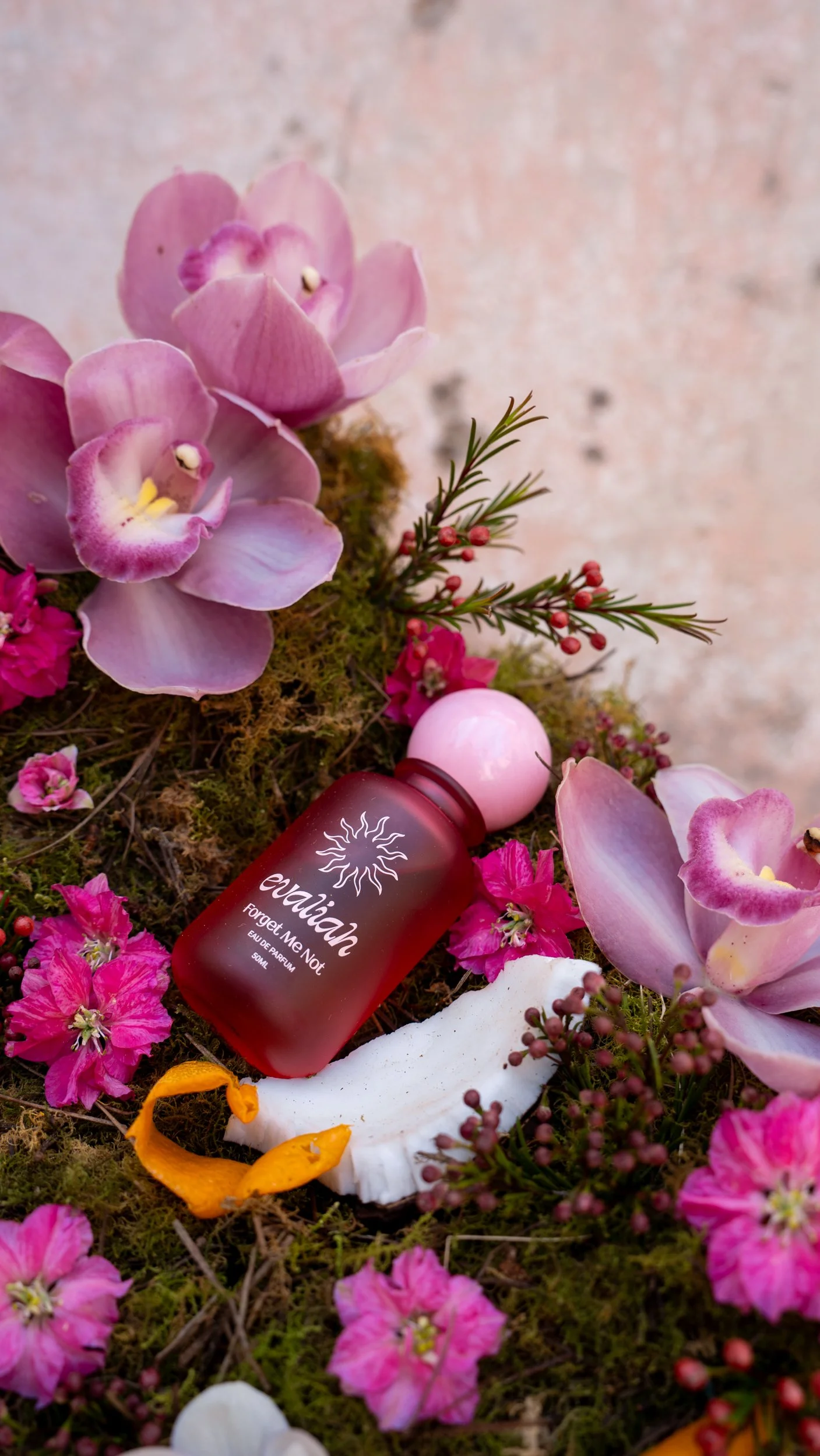 A red perfume bottle labeled 'eauihak Forget Me Not' surrounded by pink and purple flowers, moss, and small red berries, with a backdrop of a textured stone wall.