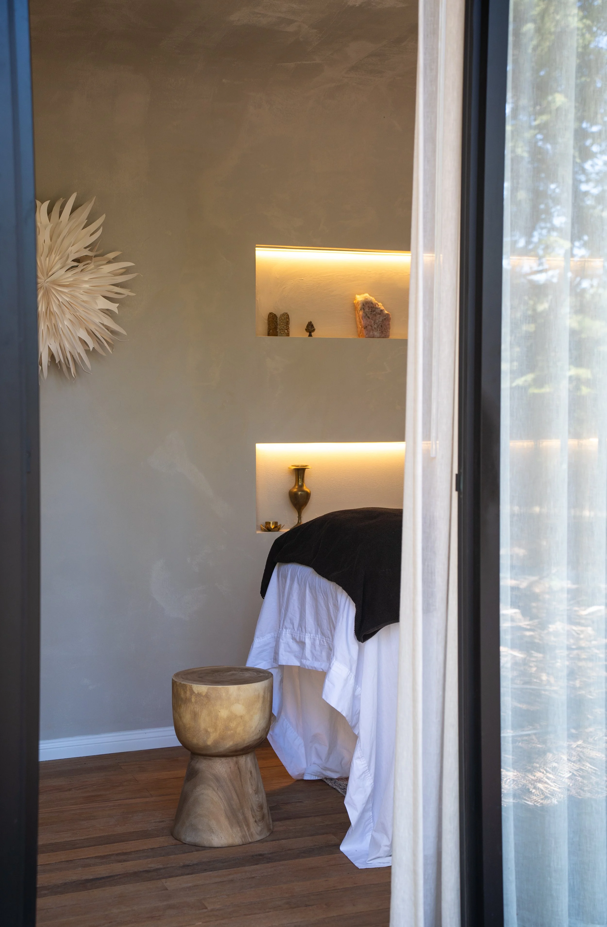 A cozy bedroom with a bed, a wooden stool, built-in wall shelves with decorative items, and a large window with sheer curtains.