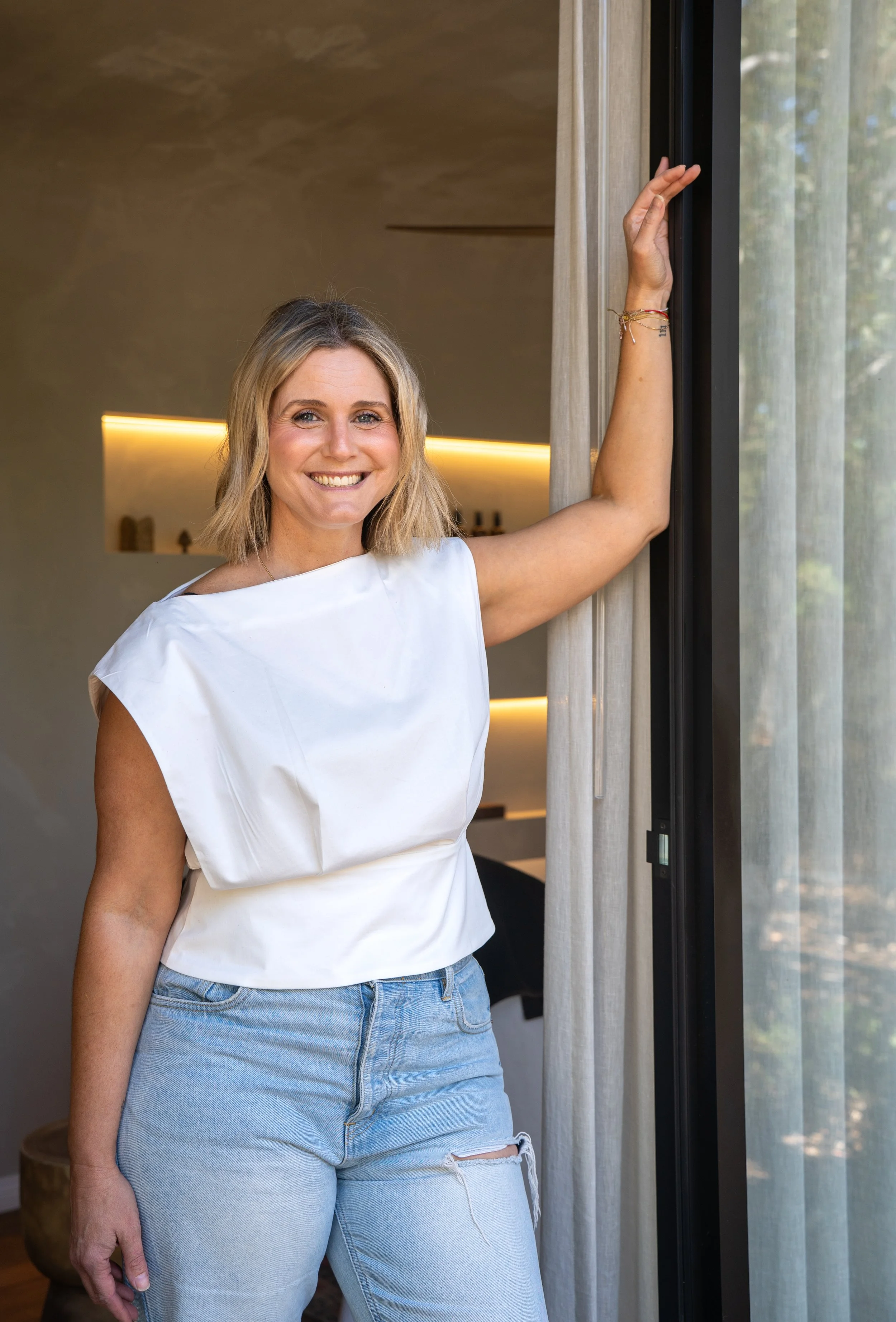 A smiling woman with blonde hair wearing a white top and light blue ripped jeans standing next to a sliding glass door. She is touching the door frame with her right hand, with a background view of trees outside.