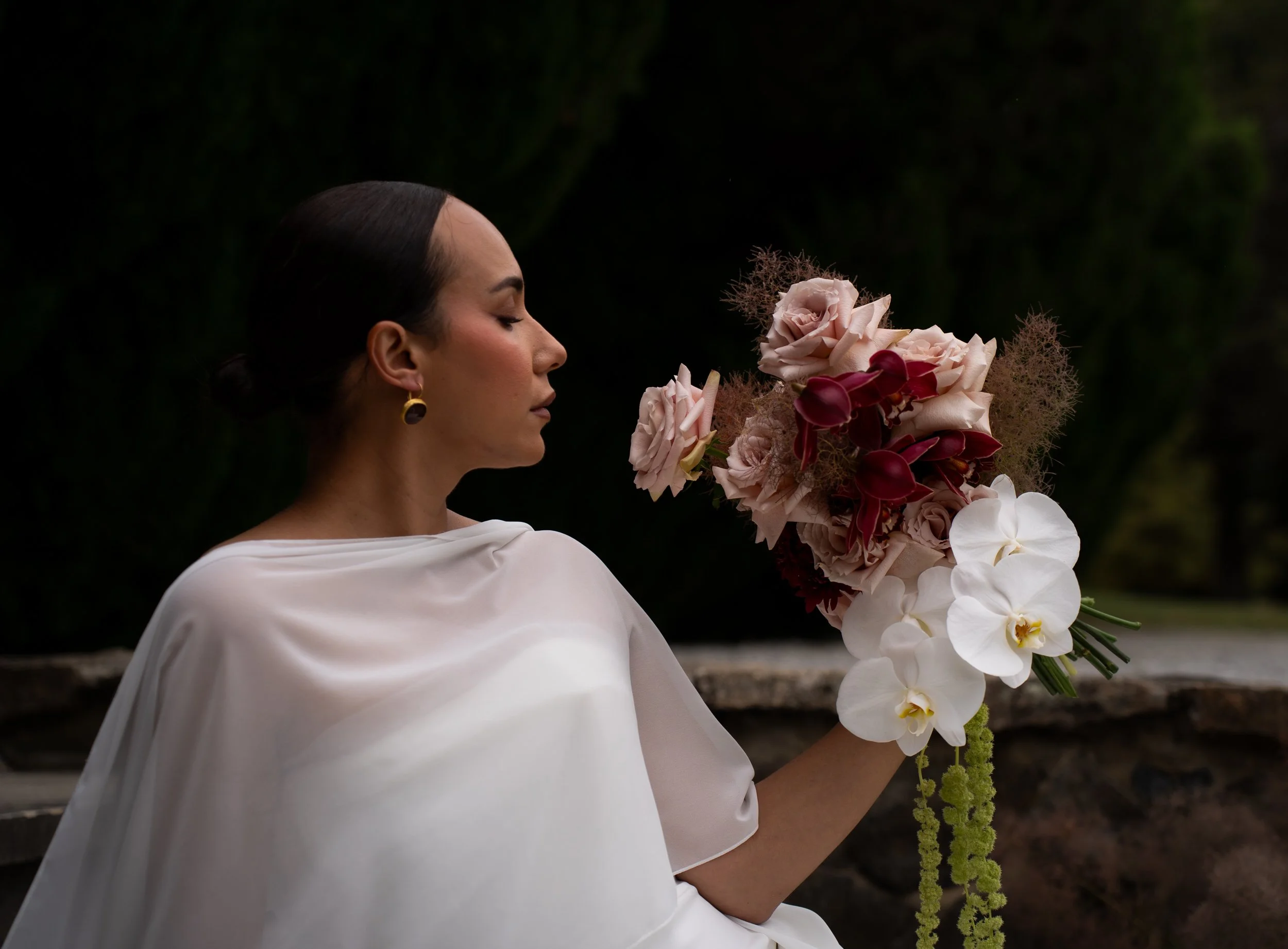 A woman with dark hair styled in a low bun, wearing earrings and a white top, is holding a large bouquet of various flowers, including orchids, roses, and other blossoms, against a dark, natural background.