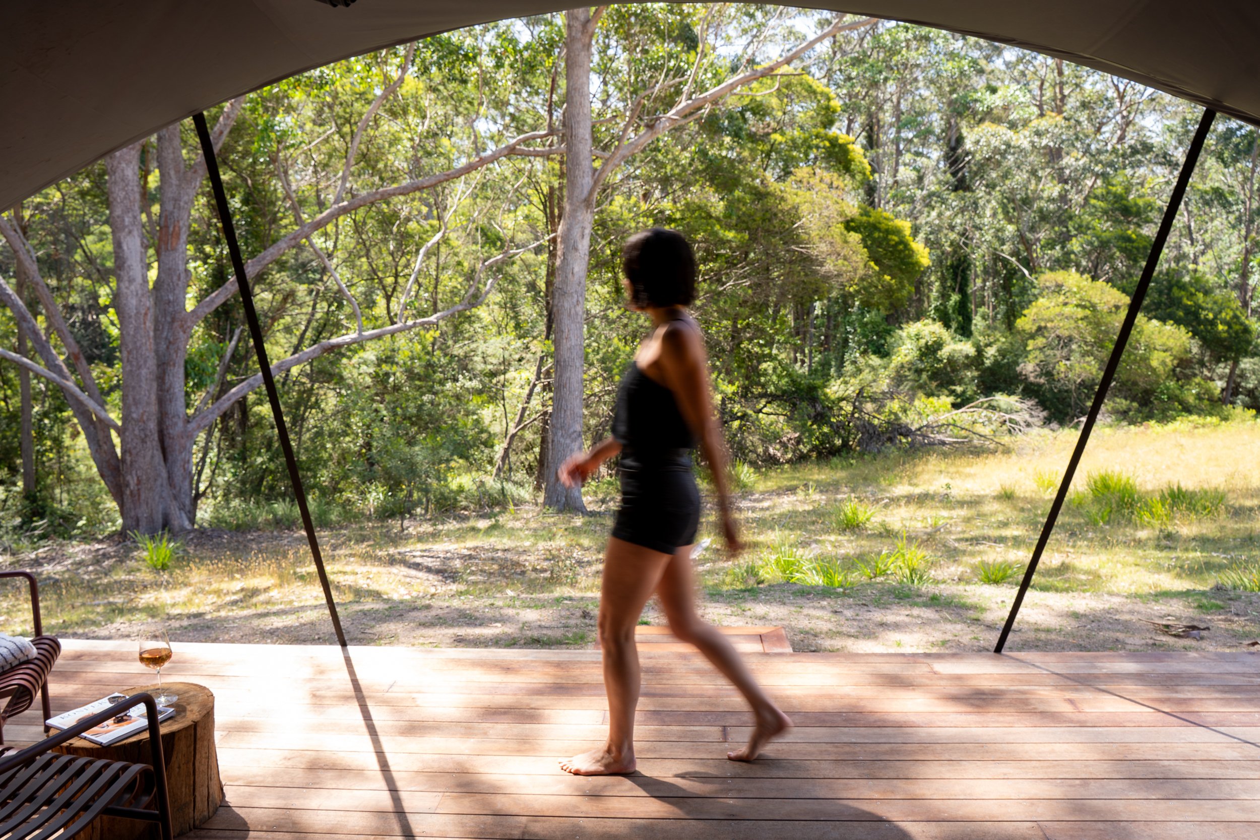 A woman in black shorts and a black tank top walking barefoot on a wooden deck, with lush green trees outside, visible through a large open canvas tent or structure.