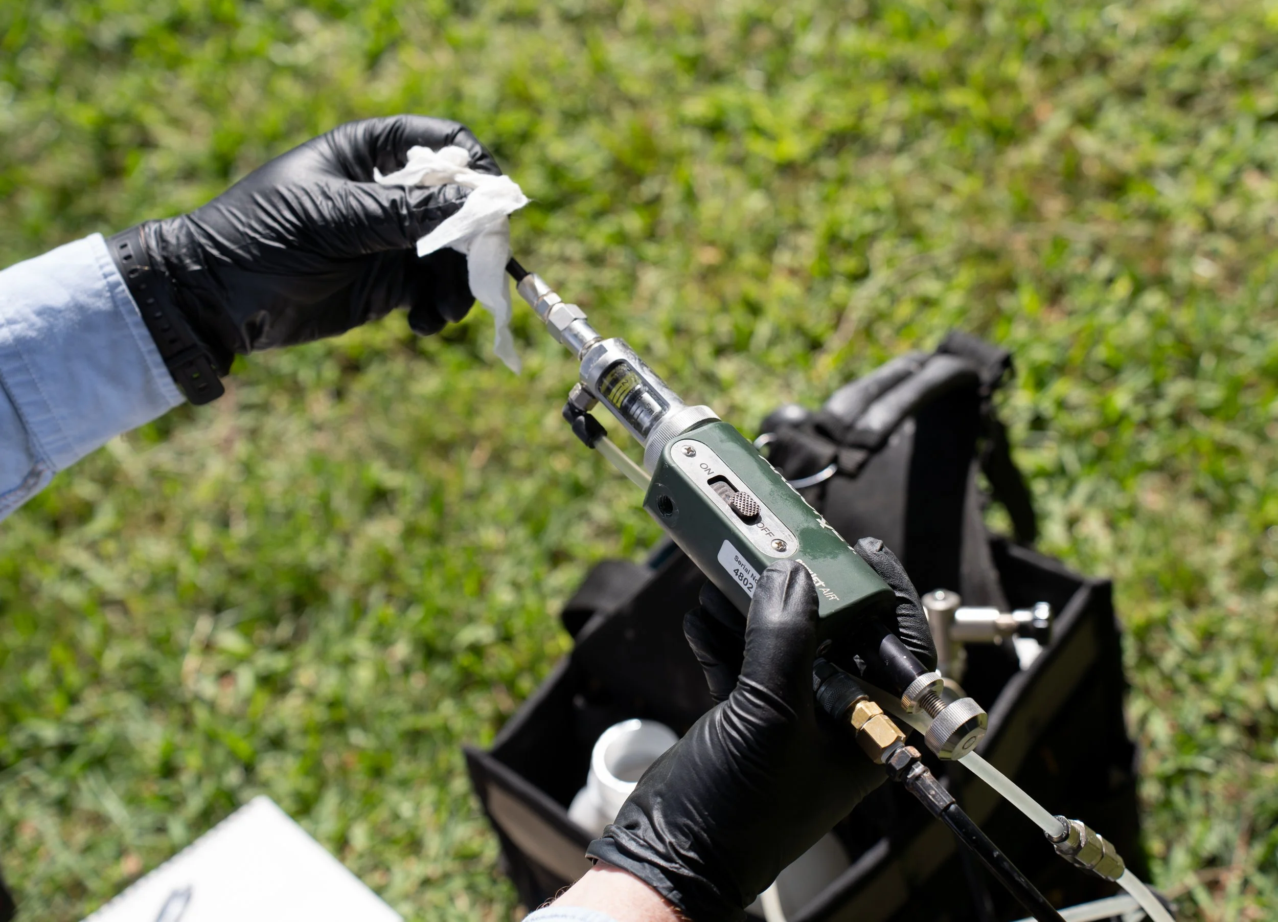 A person wearing black gloves and a light blue long-sleeve shirt holding a syringe with a nozzle, outdoors on green grass, near a black bag with various tools.