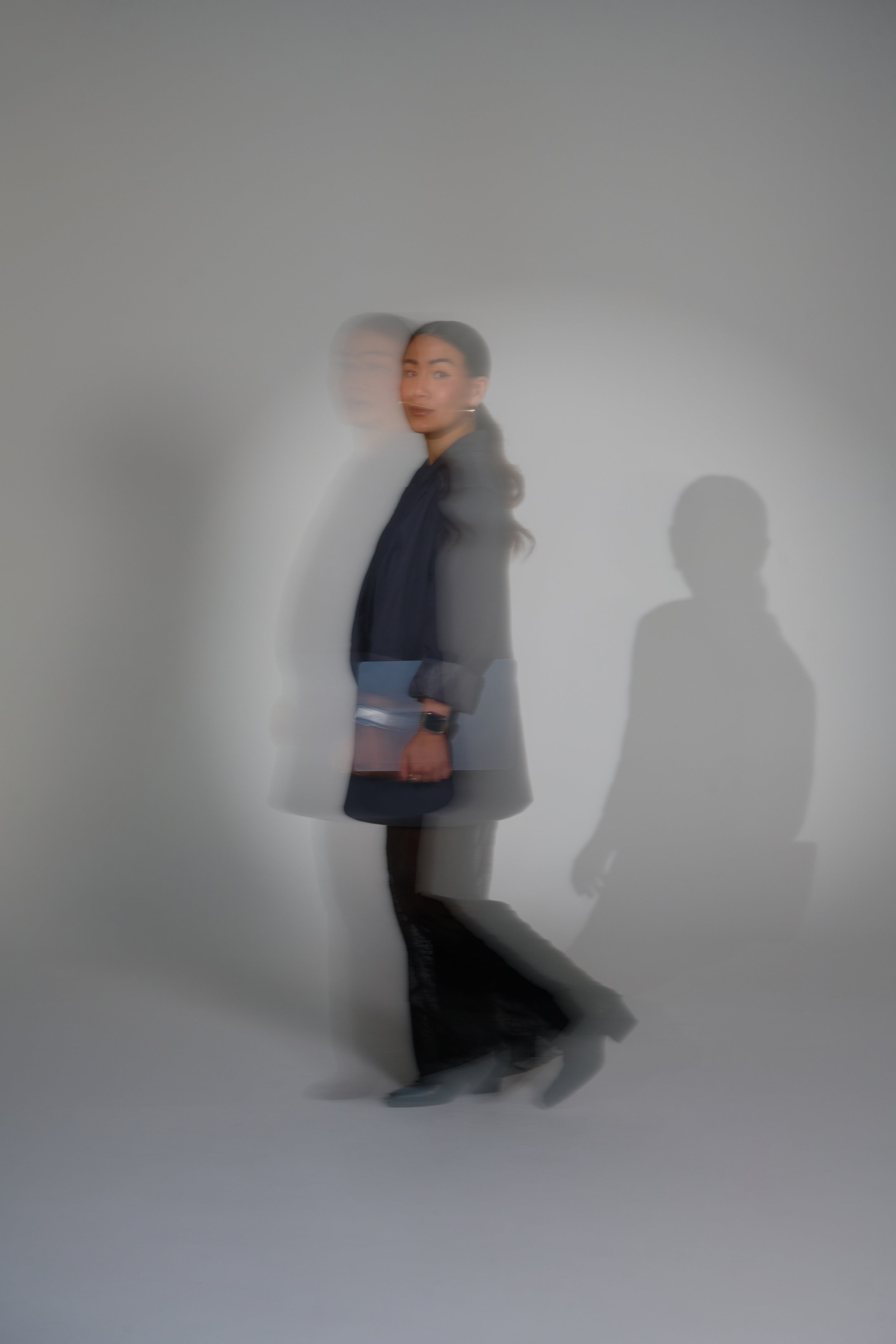 A woman standing against a plain white wall, with a motion blur effect that creates a ghostly duplicate of her. She is dressed in dark clothing and holding a notebook or folder.