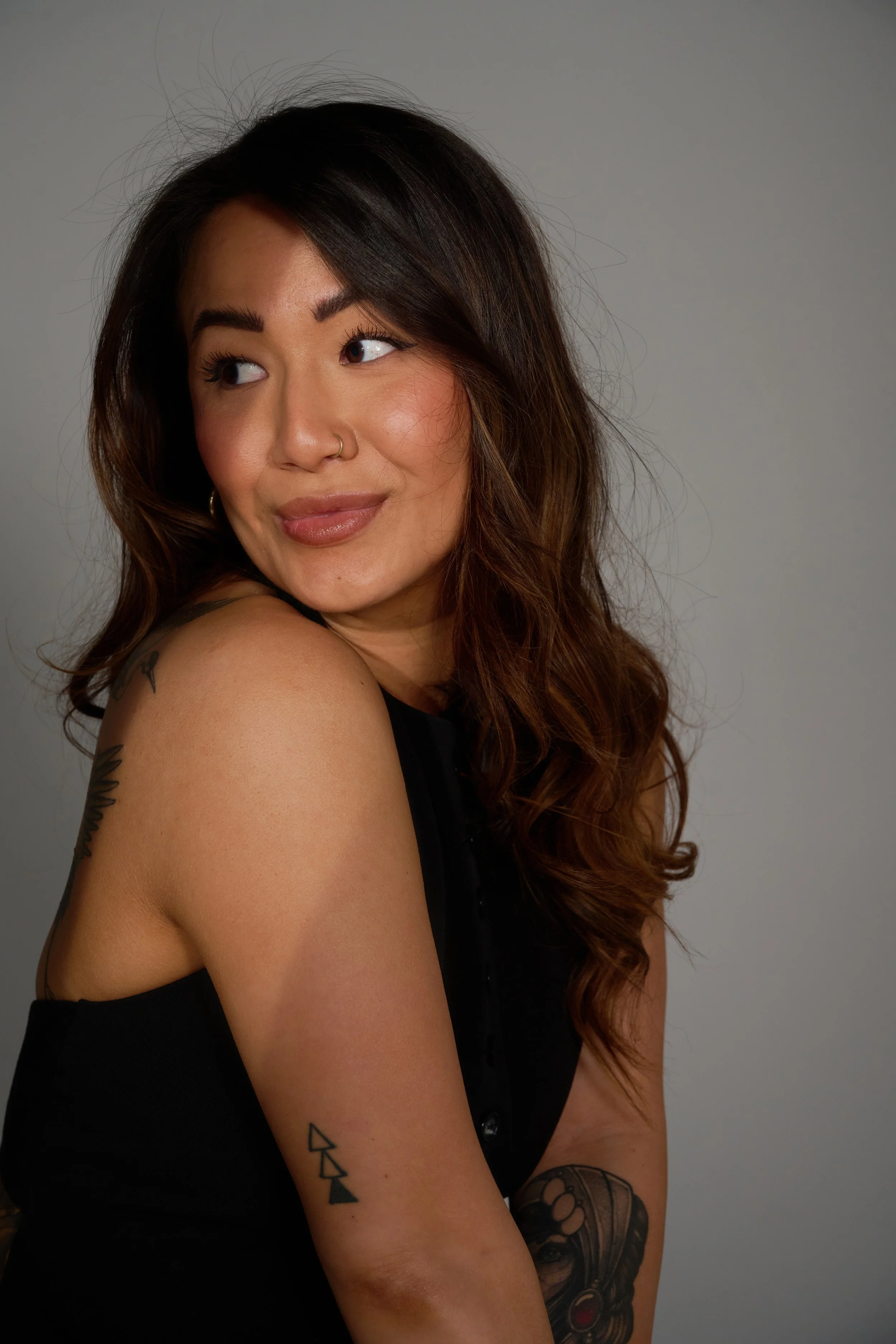 A woman with long, wavy brown hair and tattoos on her arm, looking to her left, wearing a black sleeveless top, against a plain gray background.