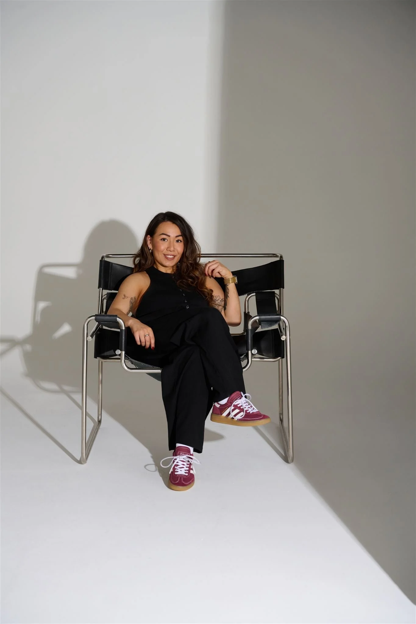 A young woman with long wavy hair, wearing a sleeveless black top, black pants, and red sneakers, sitting on a modern black chair with metal armrests. She is smiling and looking at the camera, with a shadow cast on the white and gray background.