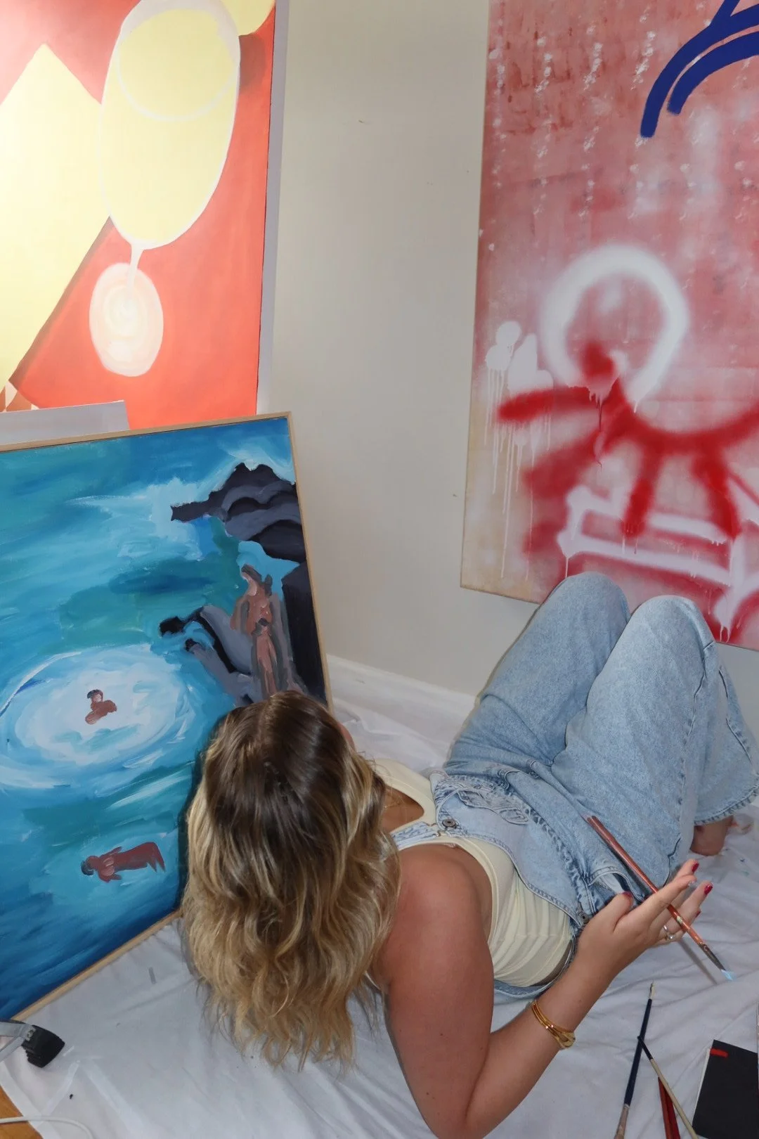 A woman with wavy blonde hair lying on the floor, holding a paintbrush, surrounded by large abstract paintings with bright colors and bold designs in an art studio.