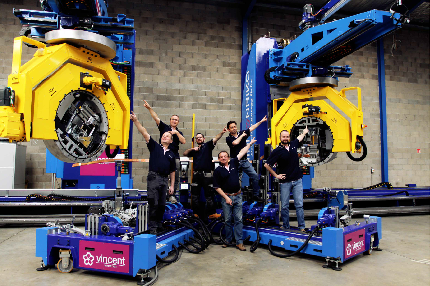 Group of five men in black shirts celebrating next to large industrial robotic machines in a warehouse or factory setting.
