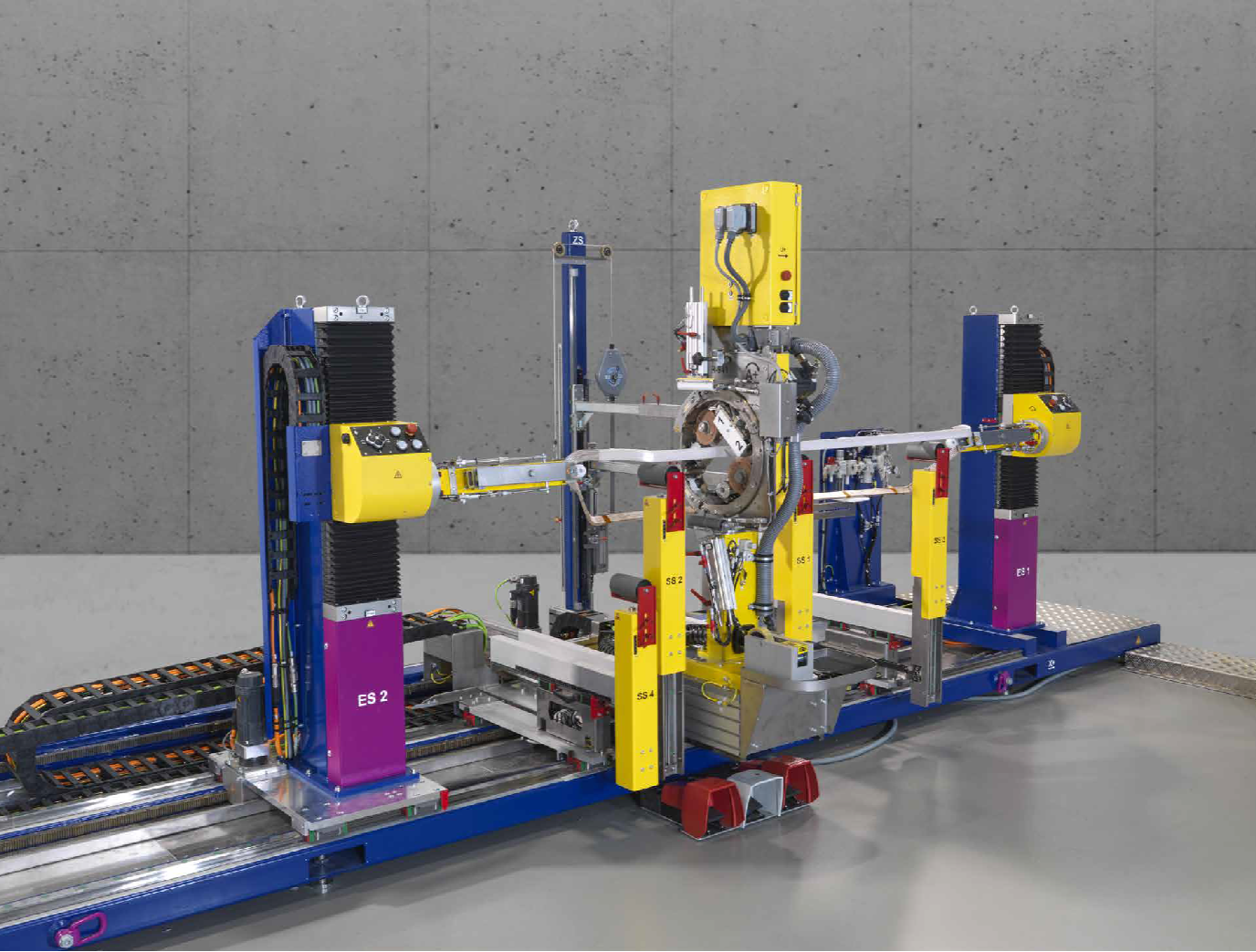 A robotic manufacturing or assembly system with multiple yellow and blue components, including robotic arms, conveyor tracks, and control panels, situated in an industrial setting with grey walls.