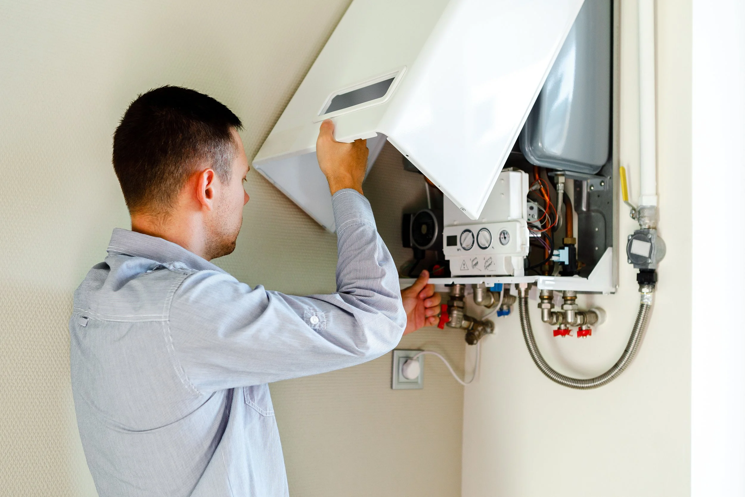 A man repairing or inspecting a wall-mounted boiler or water heater with the panel open, exposing internal components and pipes.