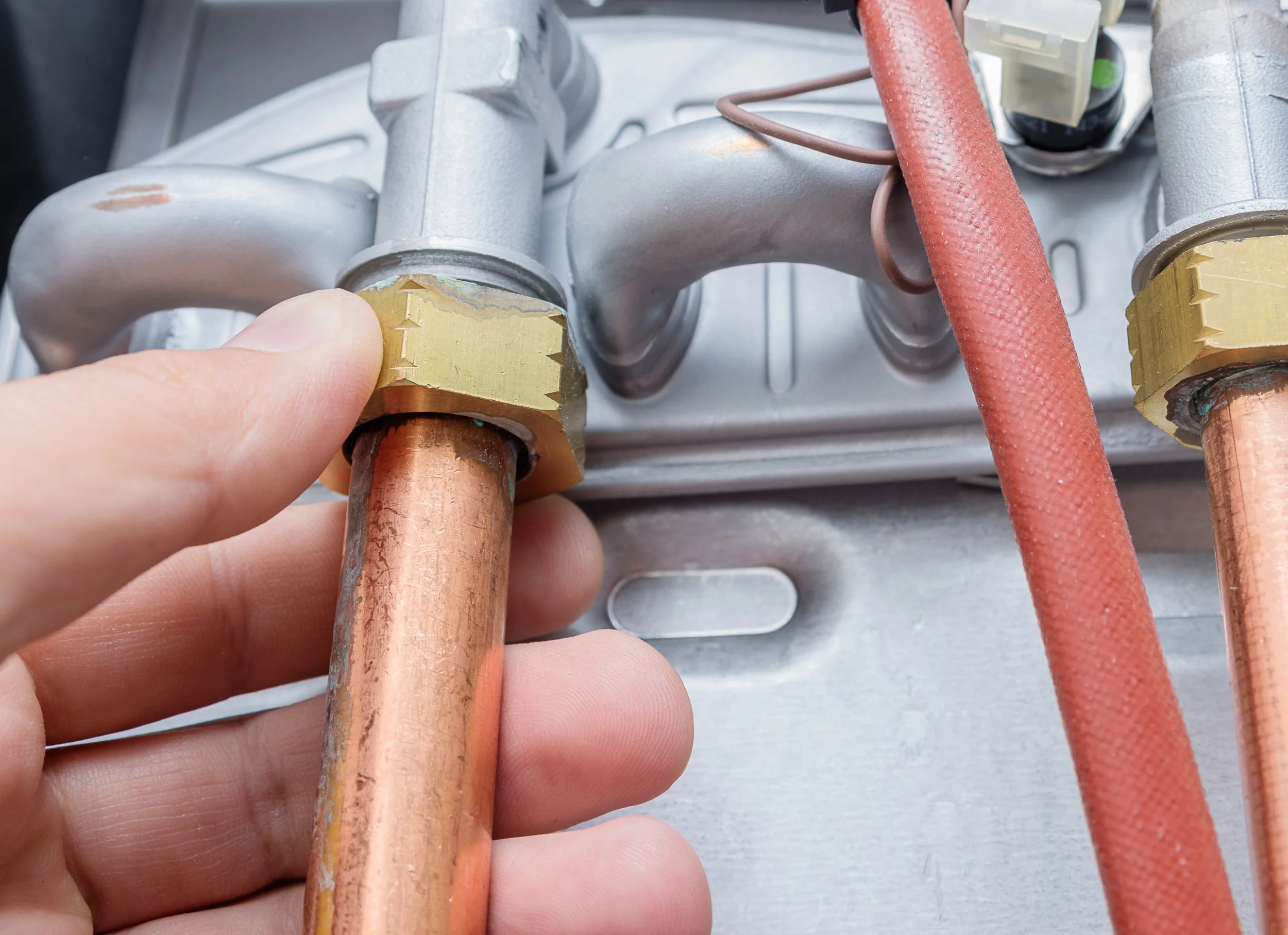 Close-up of a person holding copper piping with a brass fitting attached, part of an industrial or plumbing system. There are additional copper pipes and red insulation tubing visible in the background.