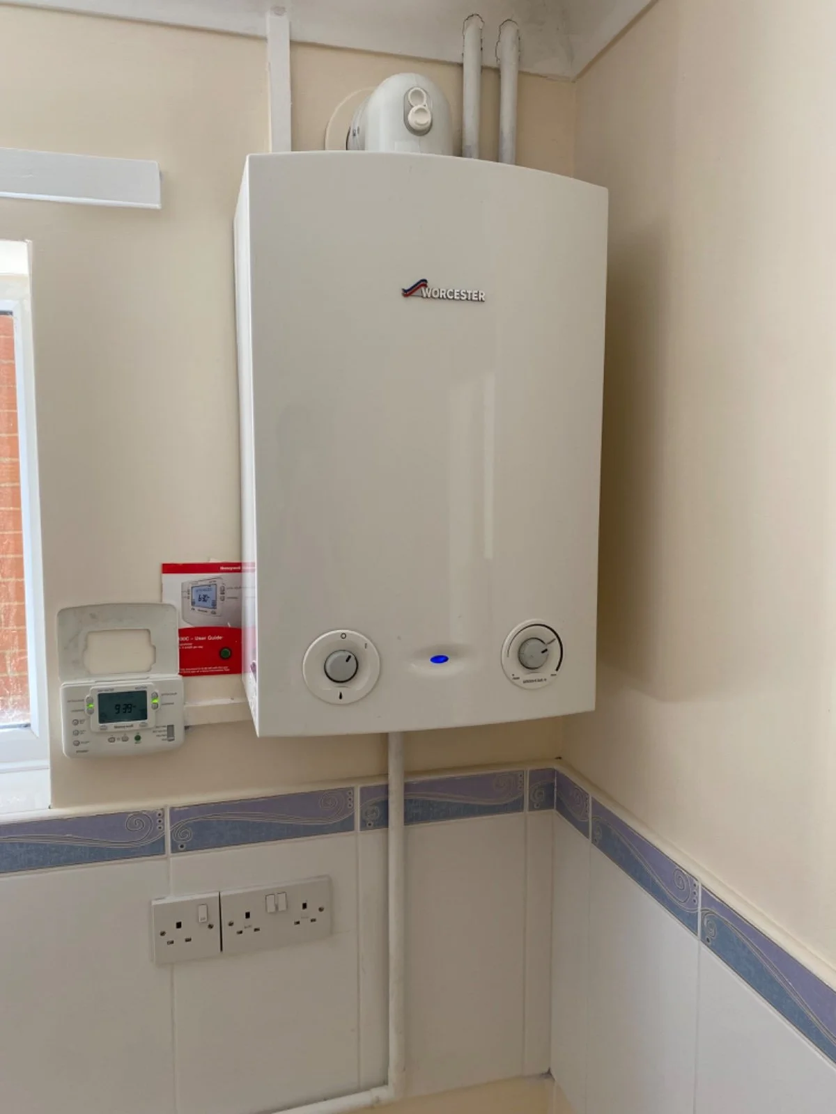 Worcester boiler mounted on a wall in a room with electrical outlets below and a small window to the left.