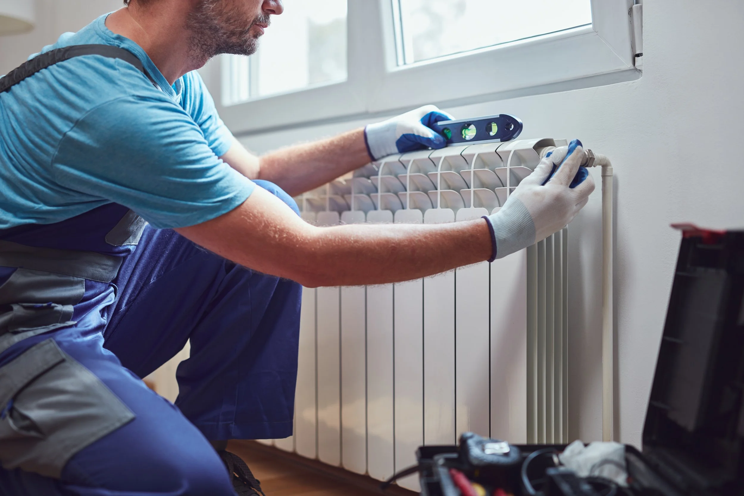 A plumber installing or repairing a radiator near a window, using a level, wearing work gloves, and kneeling on the floor with a toolbox nearby.