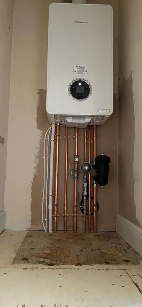 A modern tankless water heater mounted on a wall with copper pipes, a pressure gauge, and a black filter or pump below it, in a utility or laundry room. This boiler name is a Glowworm