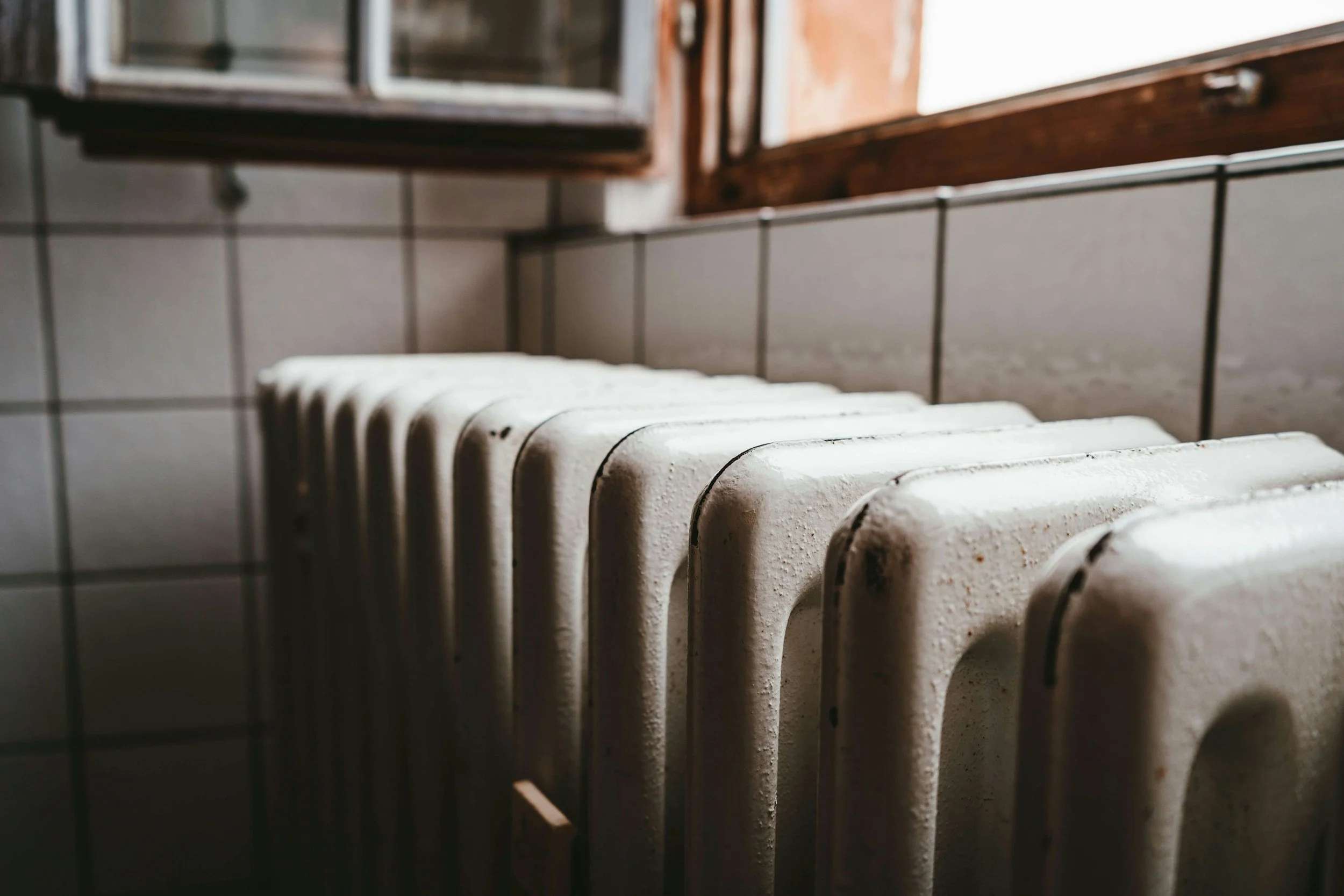 Close-up of an old white radiator next to tiled wall and wooden window frame, in a room with natural light.