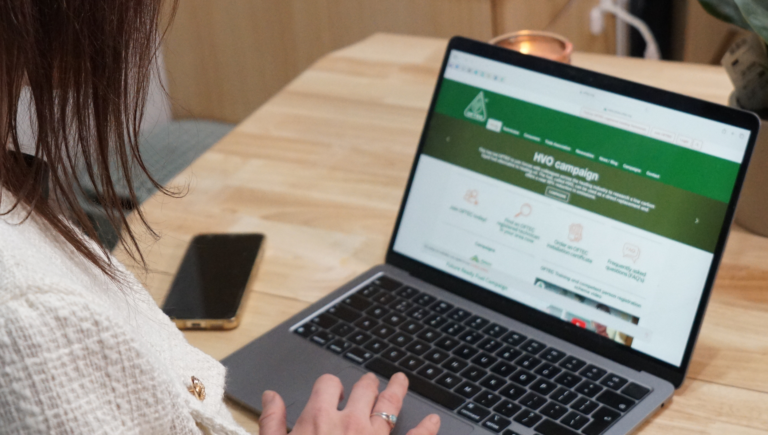 A person with brown hair wearing a white textured sweater is sitting at a wooden table using a MacBook laptop. The laptop screen displays a green website titled 'HVO campaign'. A smartphone is placed on the table next to the laptop and is turned off.