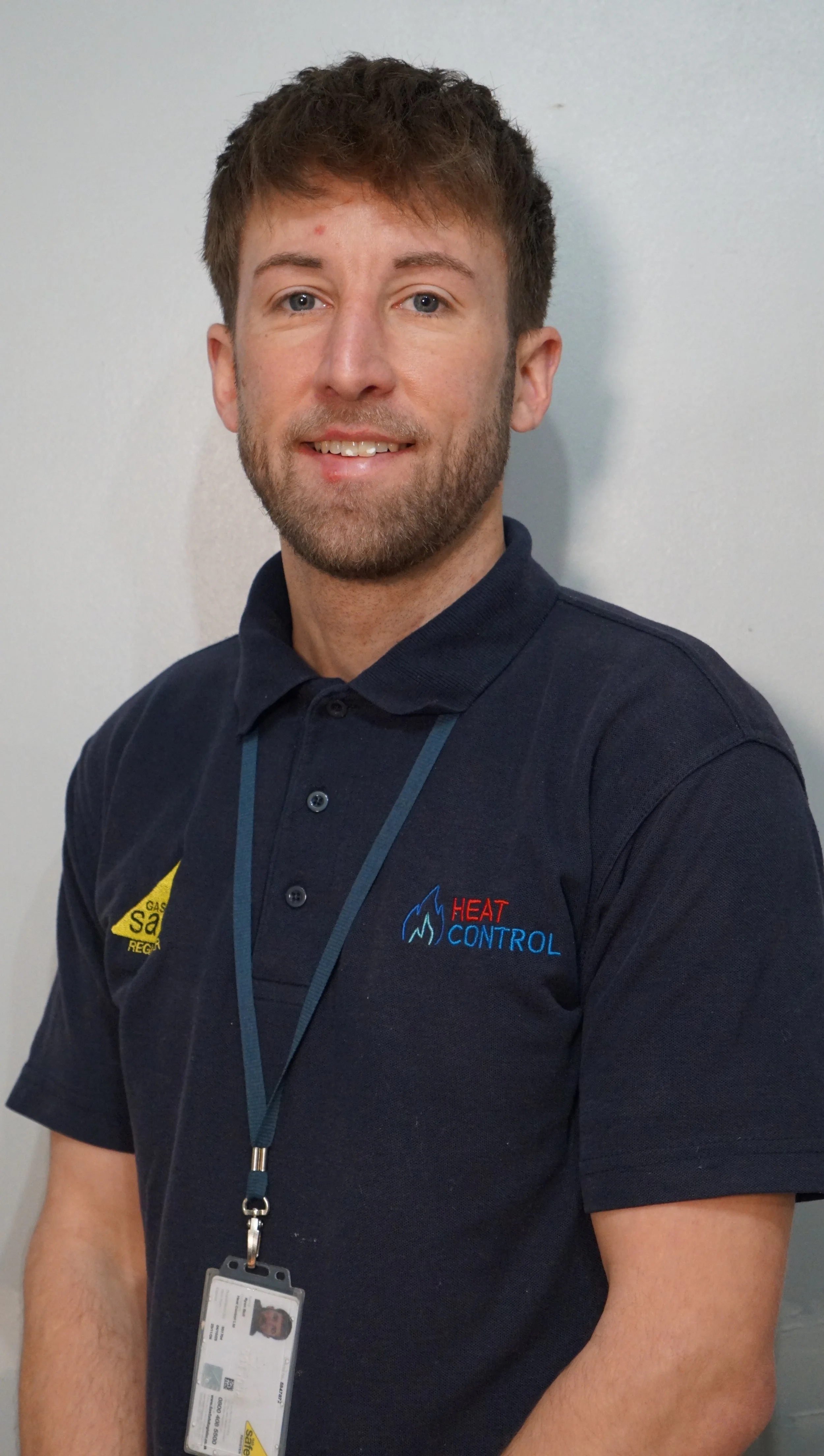 A young man with short brown hair and a beard wearing a navy blue polo shirt with a 'Heat Control' logo, standing against a white wall, with a lanyard and ID badge around his neck.