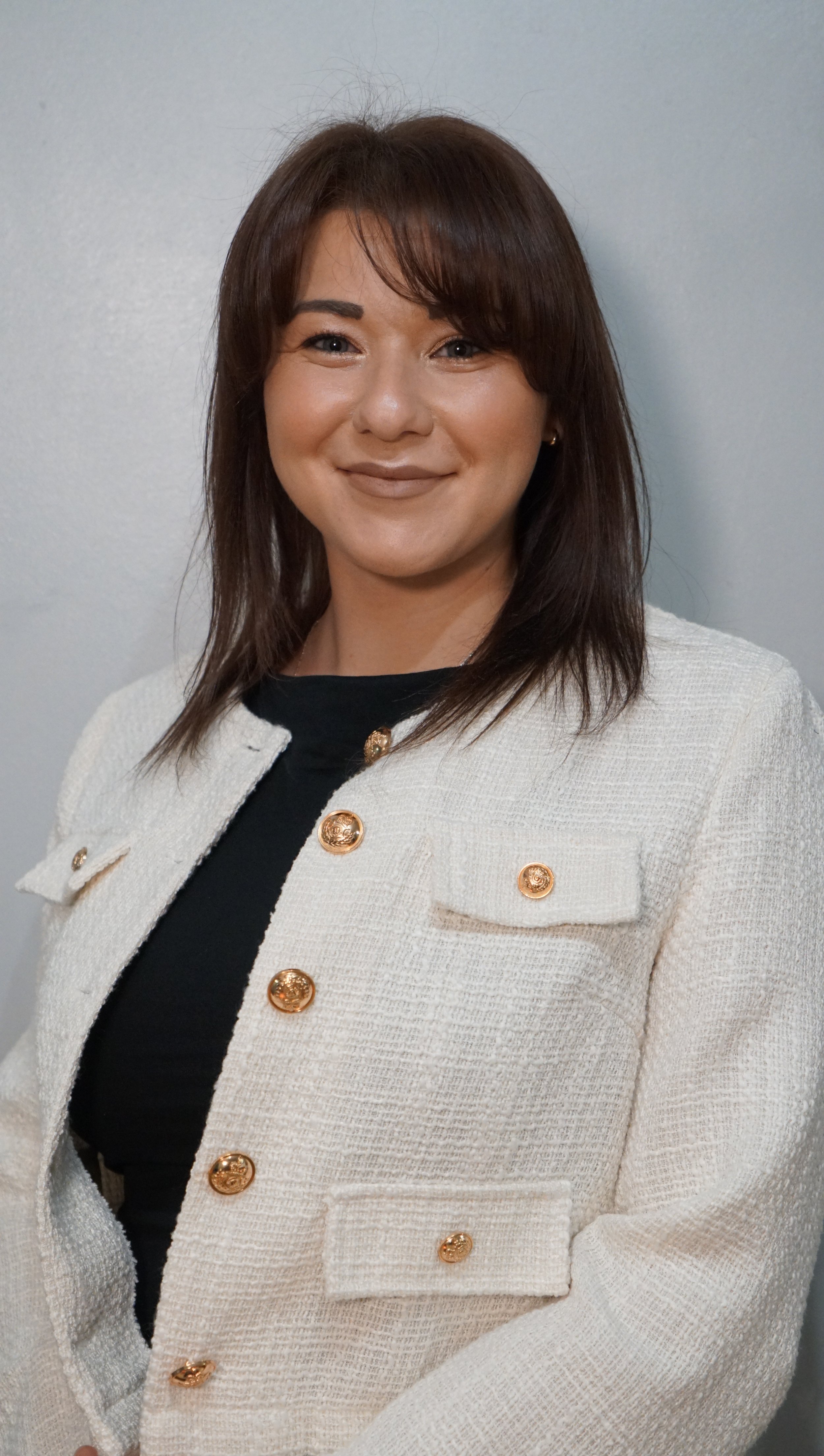 A woman with shoulder-length brown hair, wearing a cream-colored textured blazer with gold buttons over a black top, smiling at the camera against a plain light gray background.