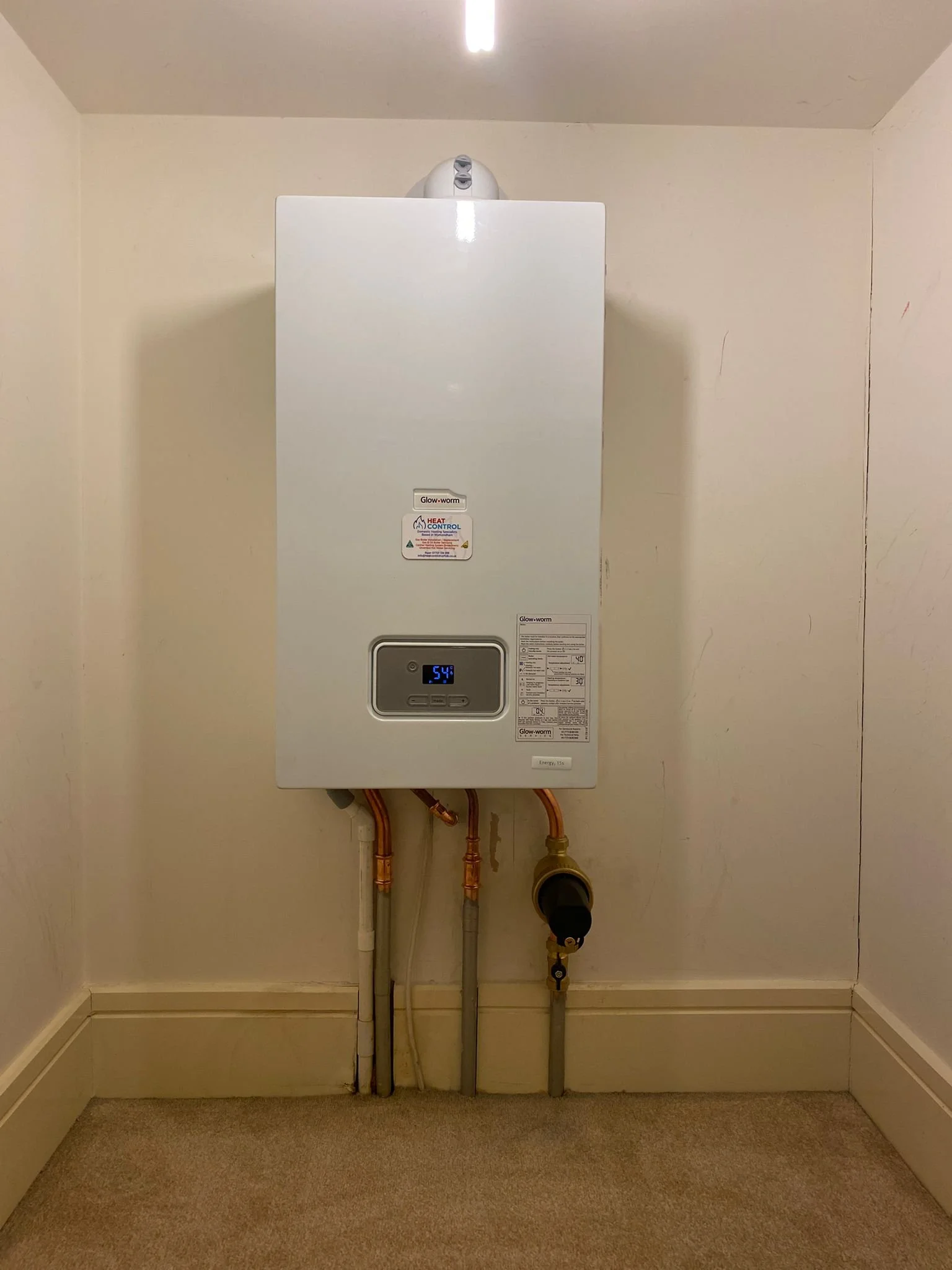 A wall-mounted white tankless water heater with digital display showing 54 degrees, pipes underneath, and a beige wall and carpet.